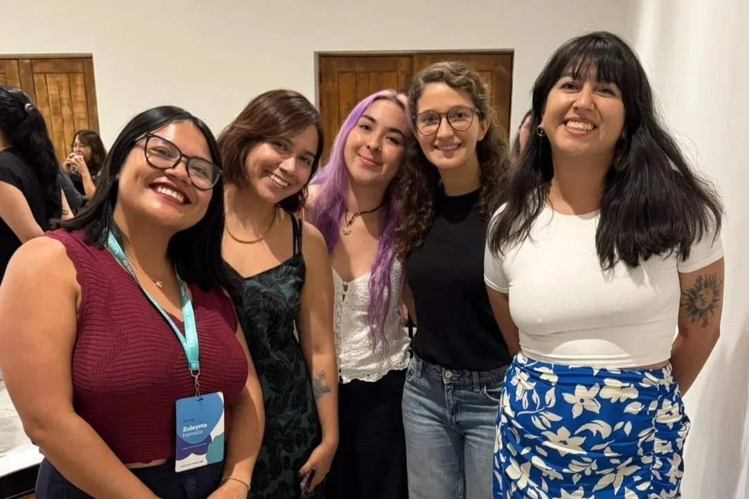 Cinco mujeres sonriendo y posando juntas en un ambiente interior, parecen estar en un evento o reunión, mostrando diversidad en estilos y edades.