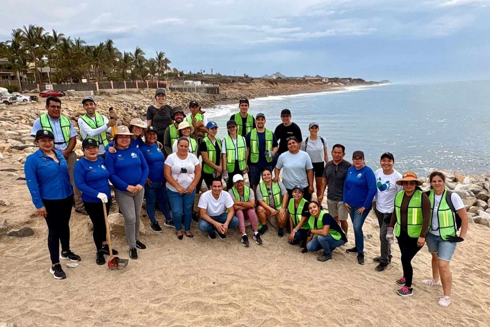 Un grupo de personas en una playa con rocas, vestido con camisetas y chalecos de colores, participando en una actividad de limpieza costera en un día nublado.