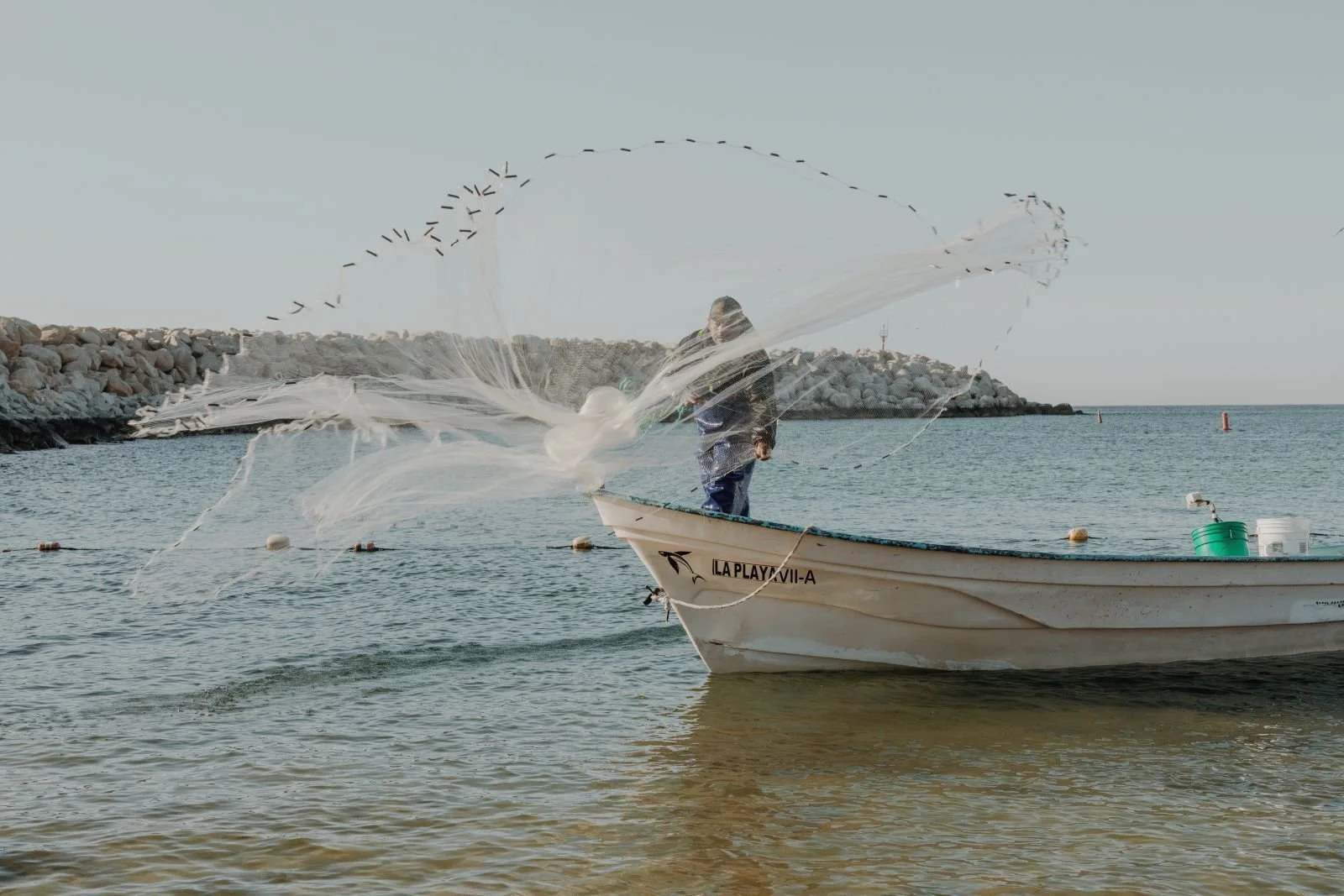 Hombre en un bote lanzando una red de pesca al mar en una playa con rompeolas y agua tranquila.