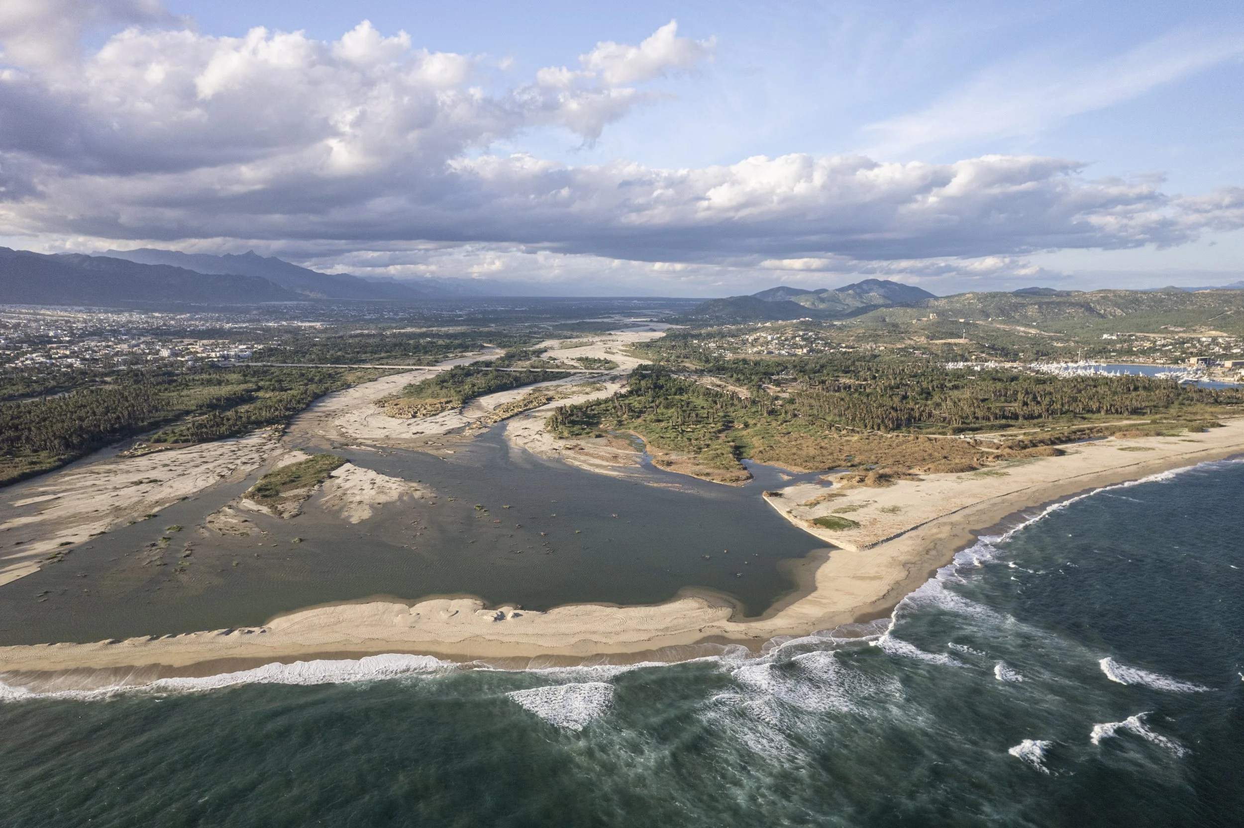 Vista panorámica de una playa de arena, agua cristalina y olas, con áreas verdes y montañas en el fondo, cielo nublado