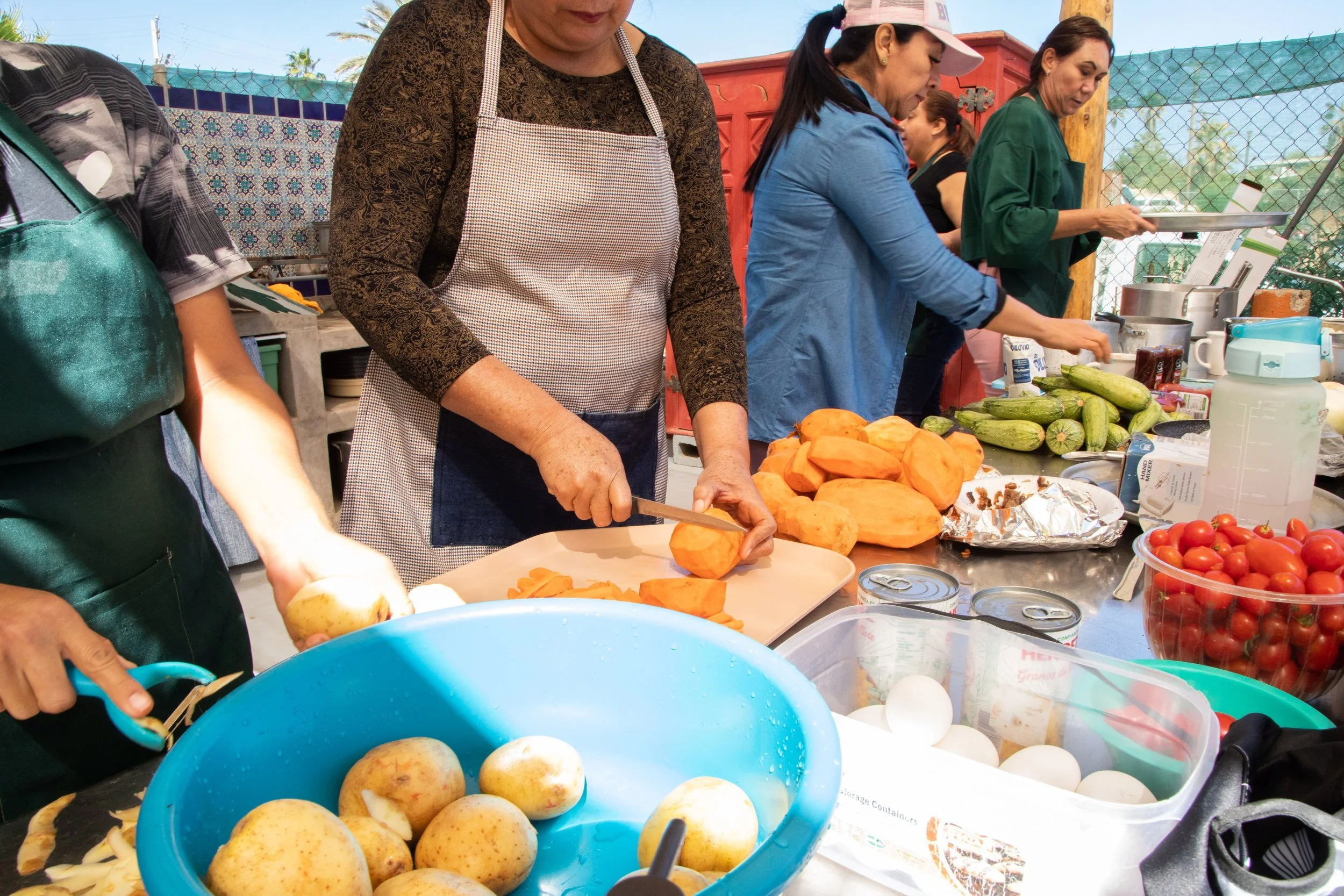 Personas cocinando y preparando ingredientes en una mesa con verduras, frutas, huevos y otros alimentos en un espacio al aire libre.