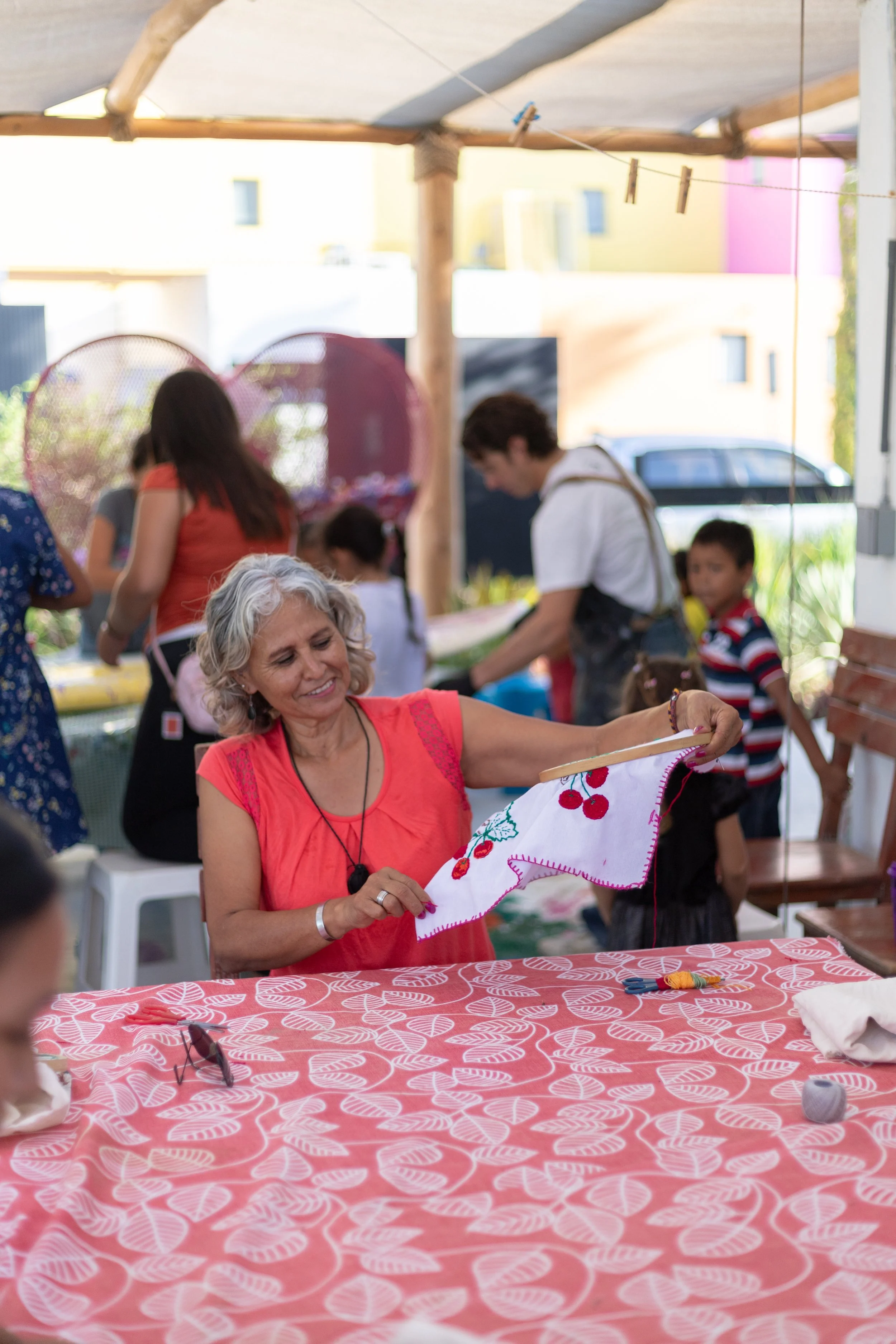 Mujer mayor sonriendo y mostrando un bordado con flores rojas y detalles en verde, en un evento al aire libre con varias personas y un toldo de sombra.