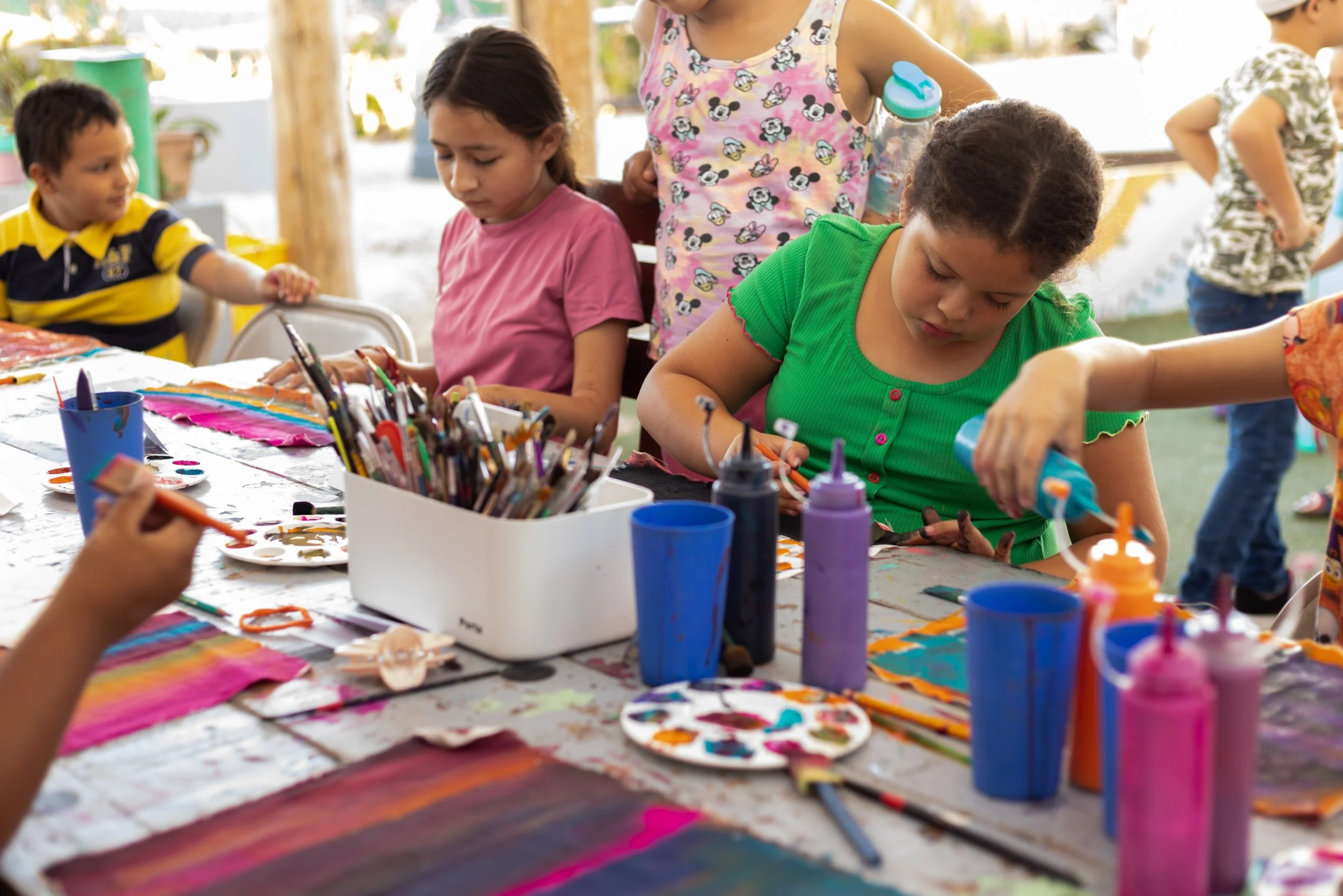 Niños y niñas en una mesa haciendo manualidades con pinturas y pinceles en un ambiente de día.
