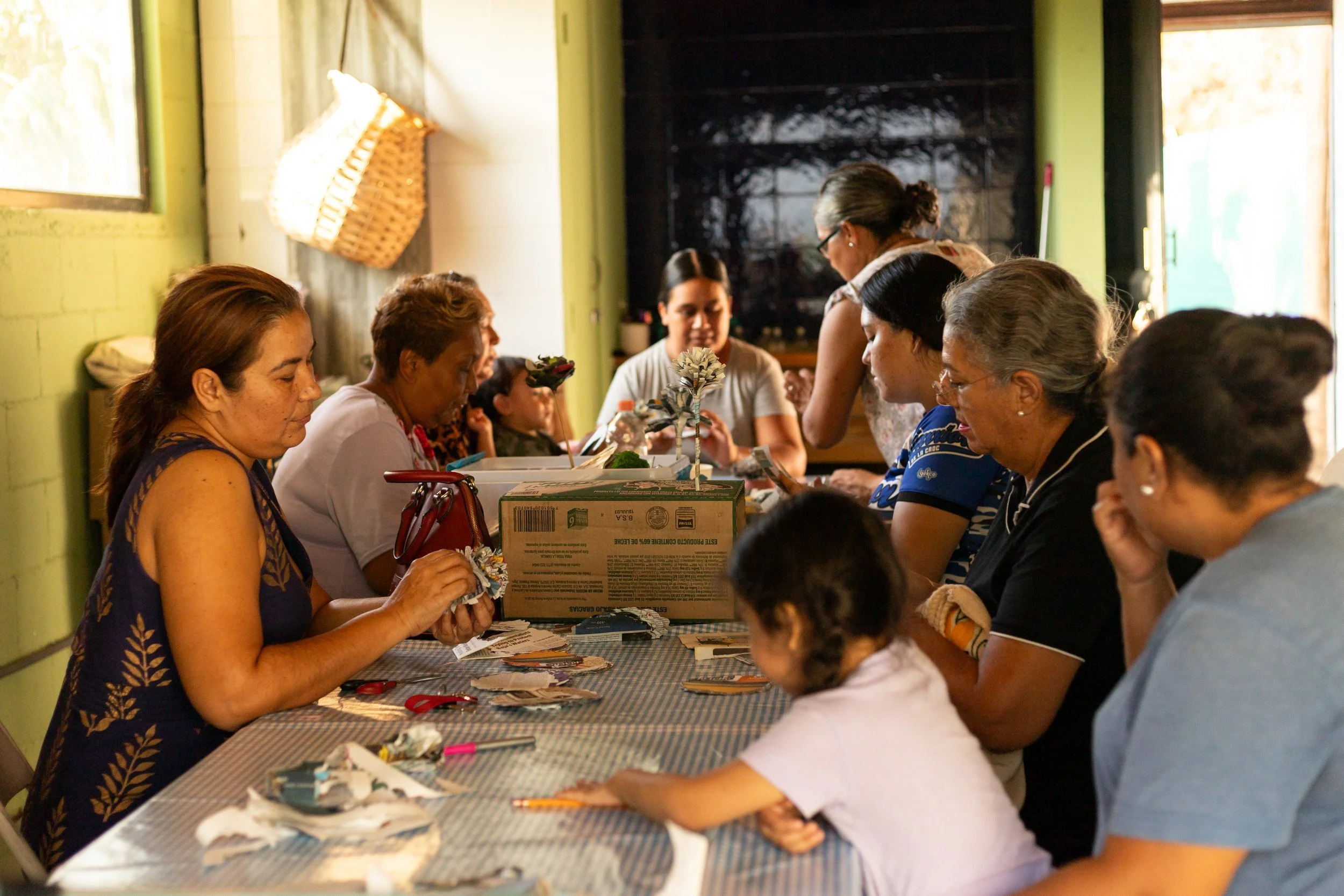 Grupo de personas en una mesa participando en una actividad de manualidades en un ambiente interior con paredes verdes y ventanas
