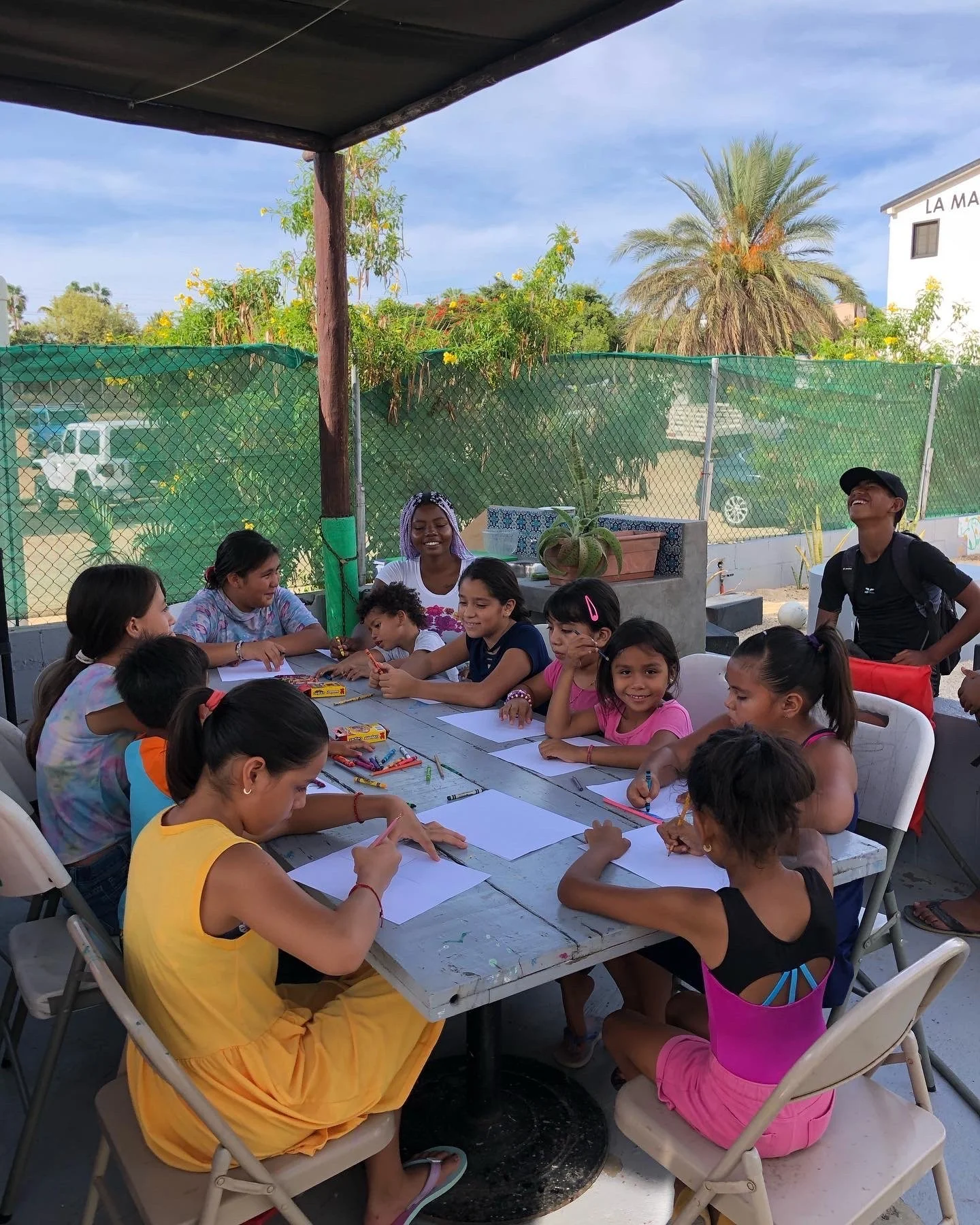 Niños y niñas realizando una actividad artística alrededor de una mesa al aire libre, bajo un toldo.
