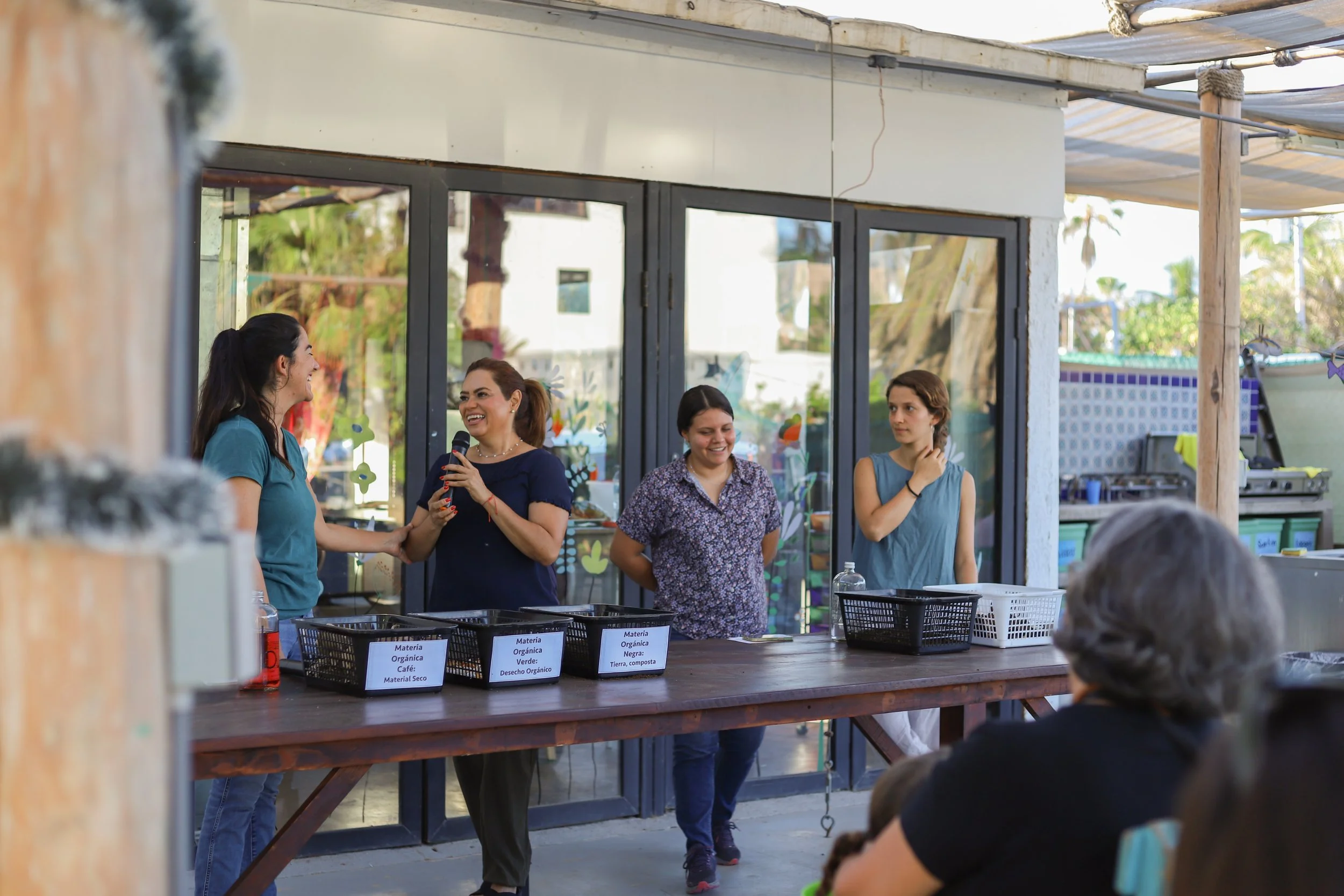 Cuatro mujeres conversando y sonriendo en un espacio abierto, con carteles en una mesa que indican diferentes tipos de materiales y residuos orgánicos. 