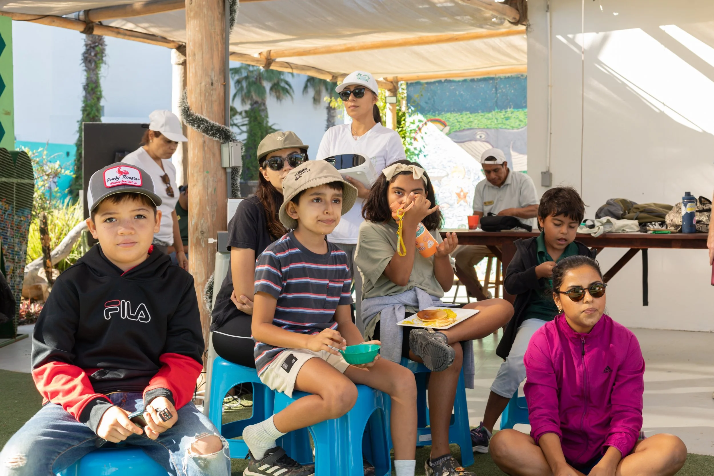 Niños y adultos en un evento al aire libre, algunos sentados en sillas de plástico, con fondo de palmeras y decoraciones coloridas.