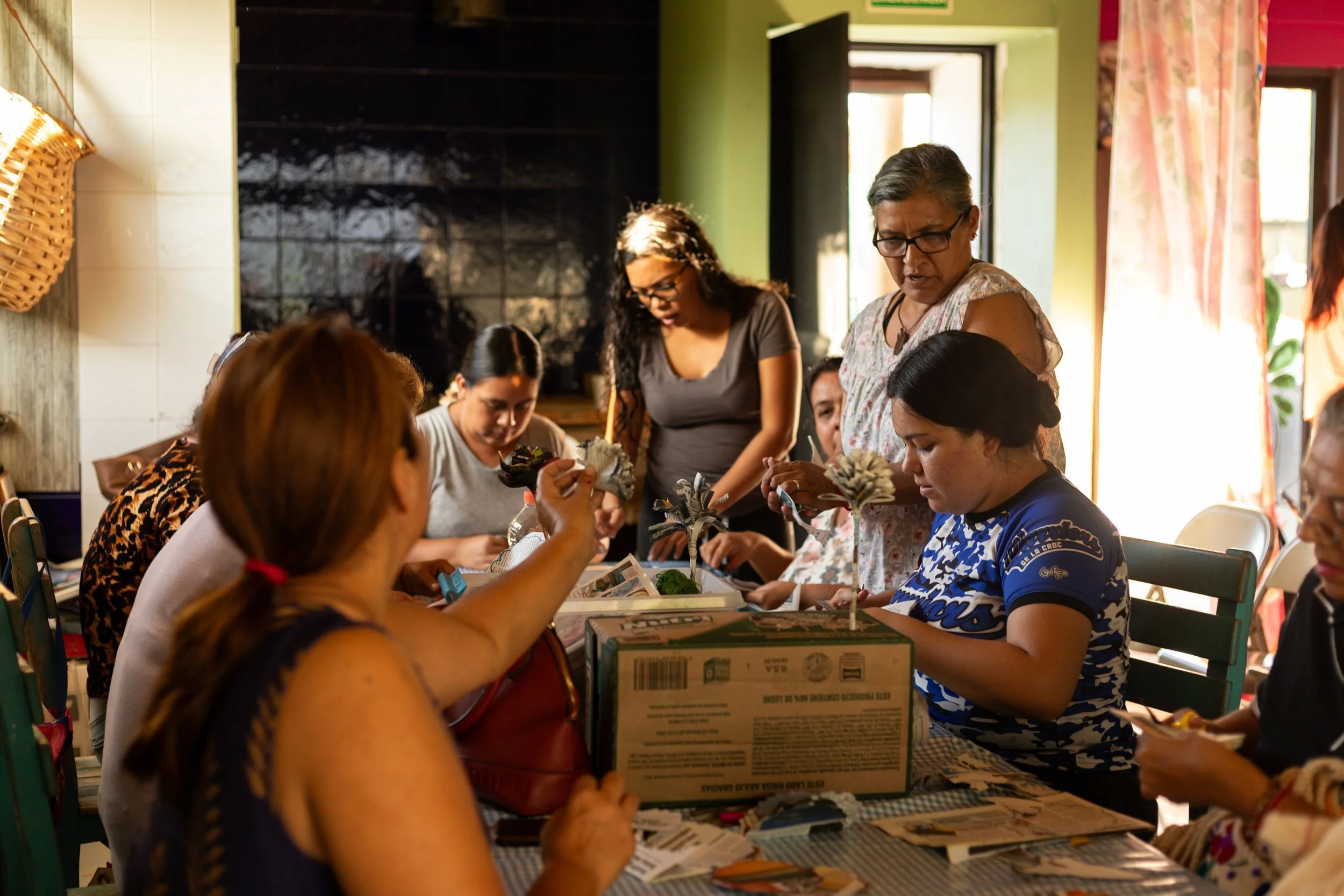 Varias mujeres y niñas trabajando manualidades en una mesa en una habitación bien iluminada y decorada con cortinas de colores.