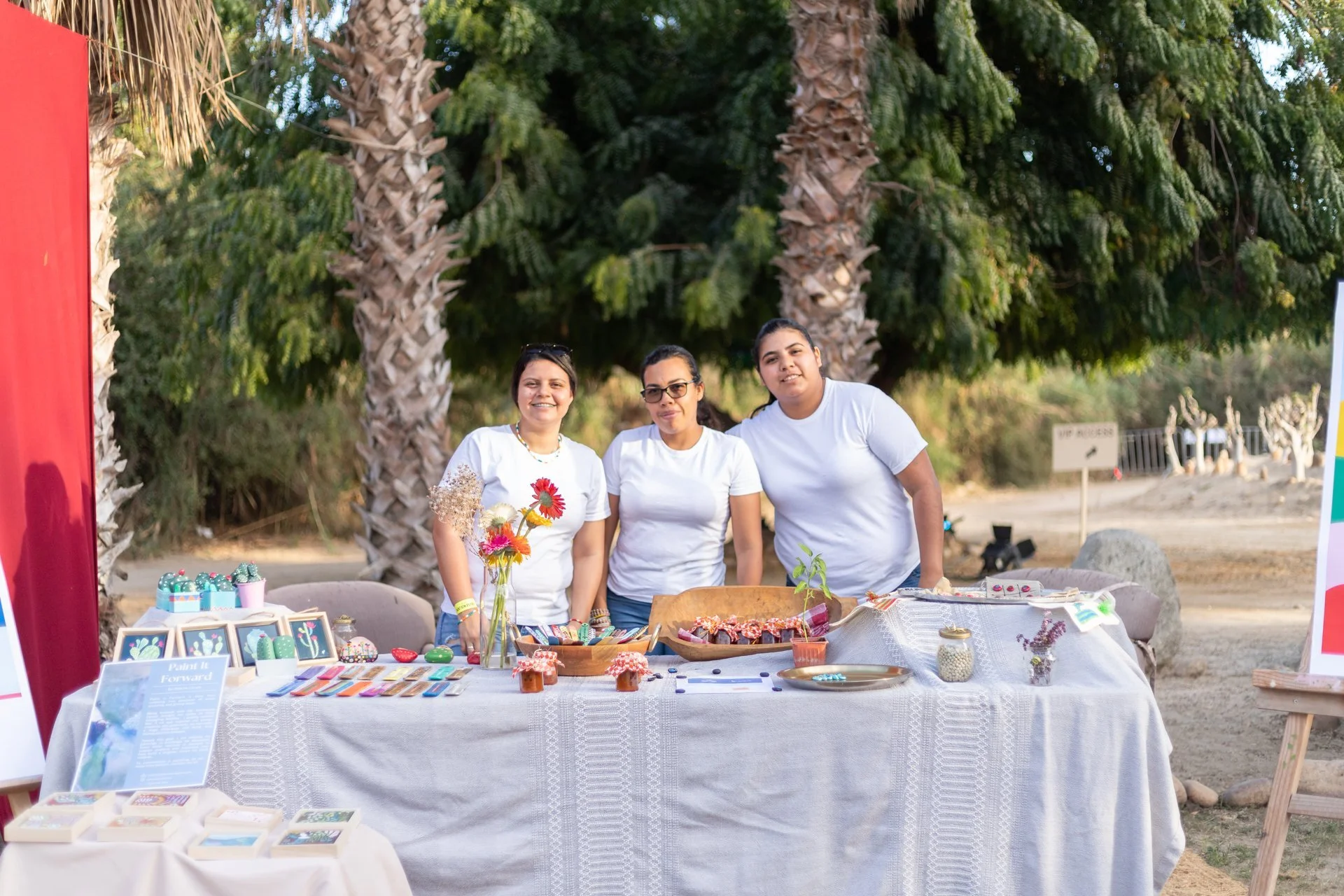 Tres personas detrás de una mesa con diversos objetos al aire libre, con árboles y palmeras al fondo.