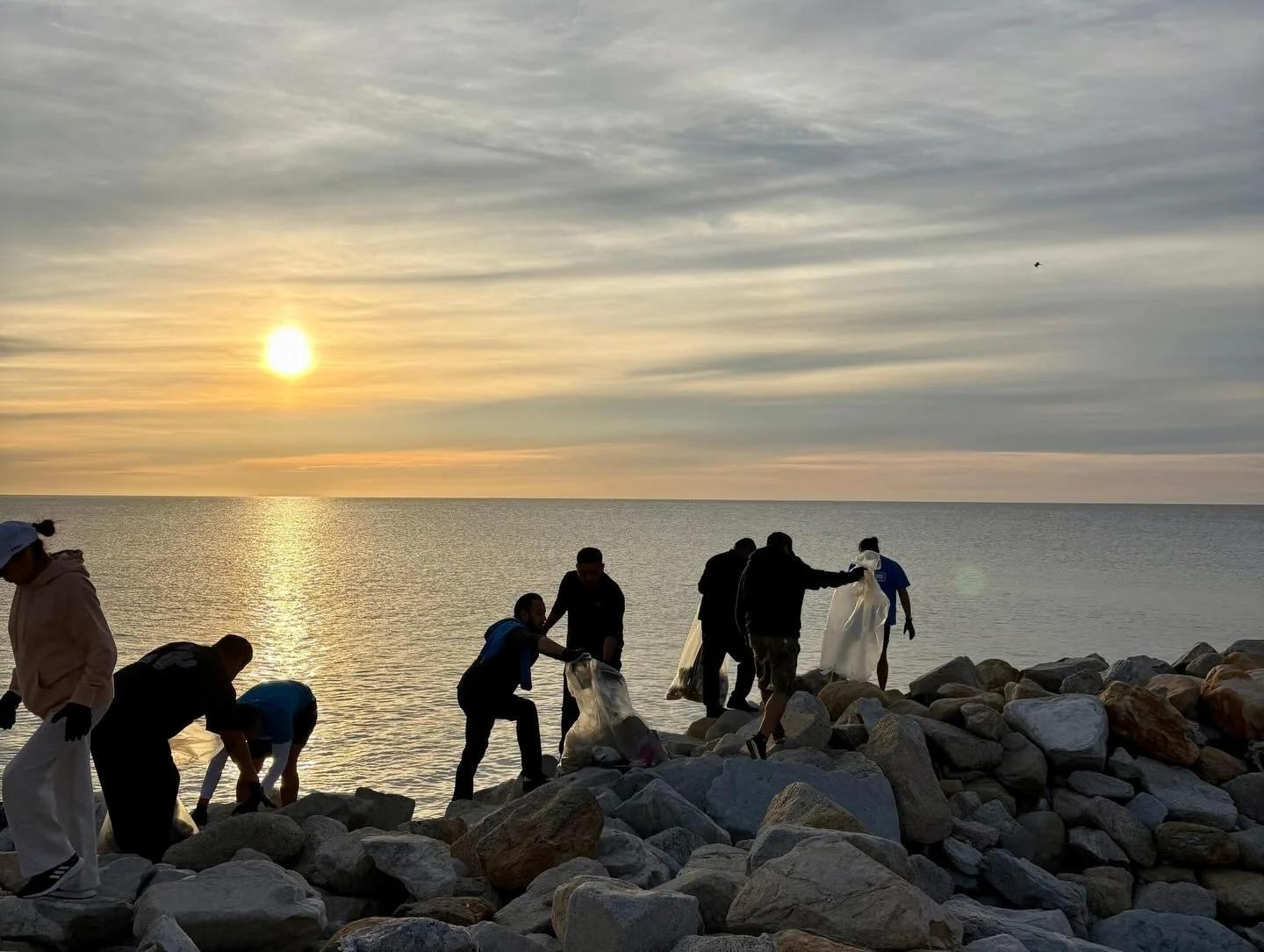 Varias personas recogiendo basura en una orilla rocosa al atardecer del mar.