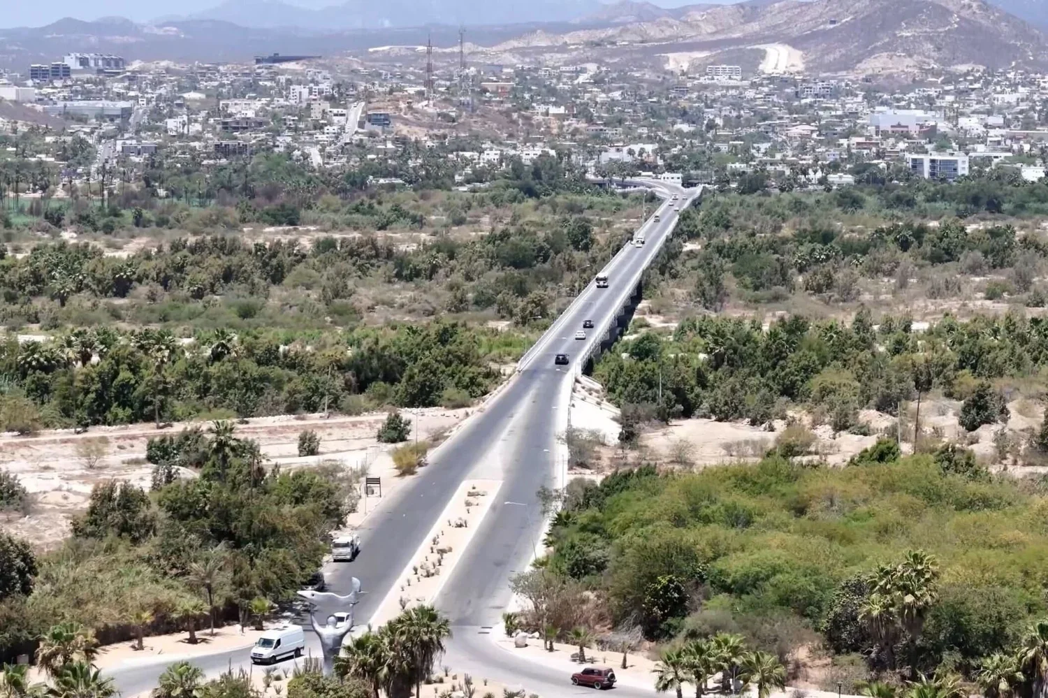 Vista aérea de una carretera con varios autos que atraviesa un área de vegetación y montañas en la distancia, con una ciudad en el fondo.