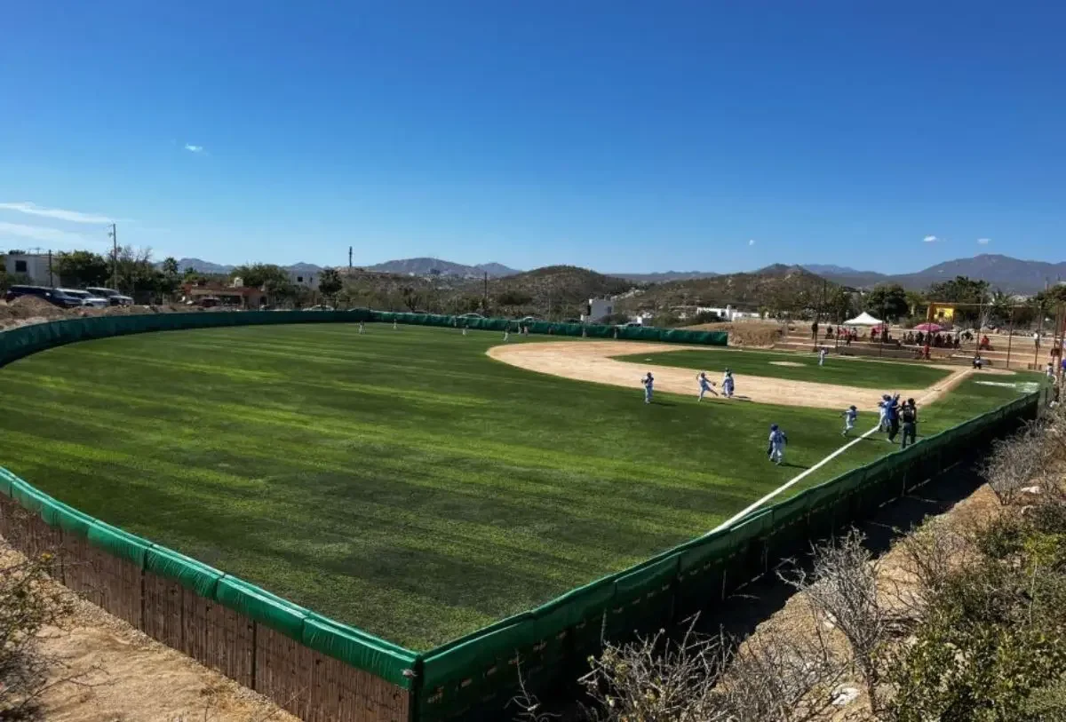 Campo de béisbol con césped, jugadores y montañas al fondo bajo un cielo despejado.
