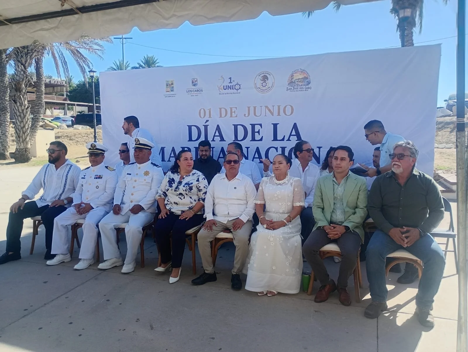Reunión bajo una carpa con varias personas sentadas en sillas, frente a un cartel que dice '01 de Junio Día de la Marina Nacional'.