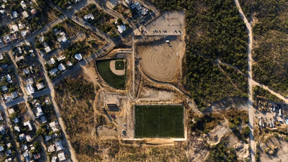 Imagen aérea de un área recreativa con campo de fútbol, pista de béisbol, parque y zonas verdes, rodeada por casas y caminos.