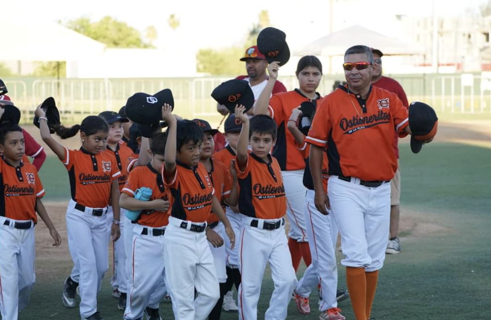 Grupo de niños y adultos en uniforme de béisbol, caminando por un campo de béisbol, algunos levantando sus gorras.