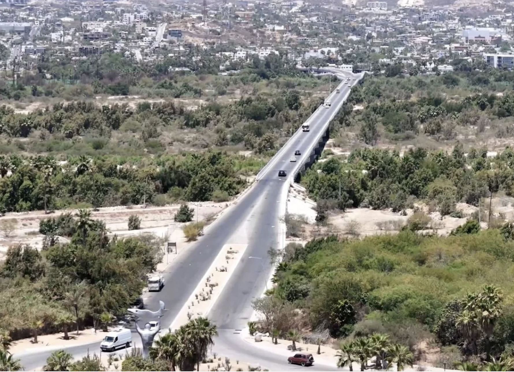 Vista aérea de una autopista elevada con varios vehículos que cruza un área de vegetación y terrenos áridos, con una zona urbana en el fondo.