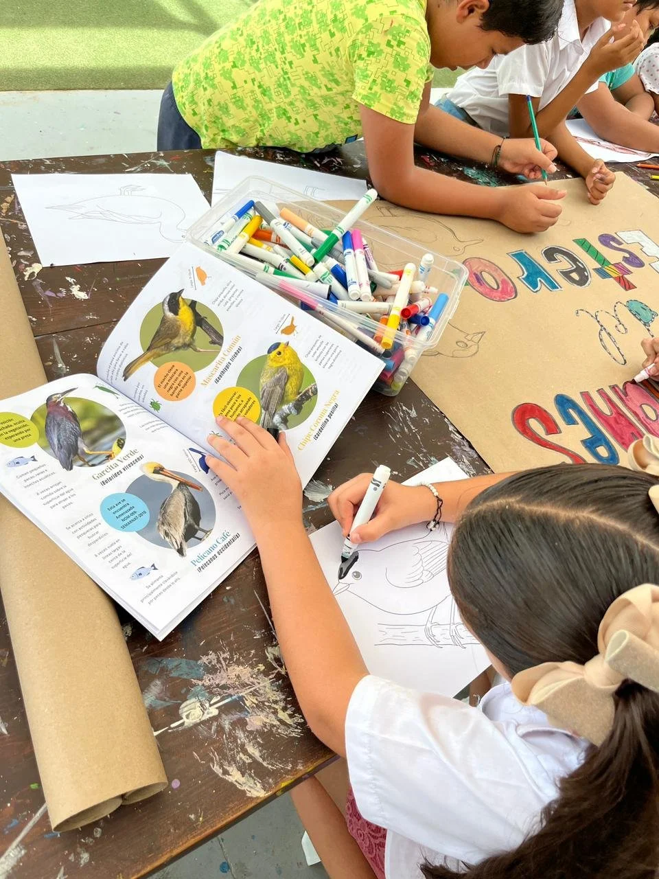 Niños pintando y leyendo sobre aves en una mesa con libros, papeles y marcadores.