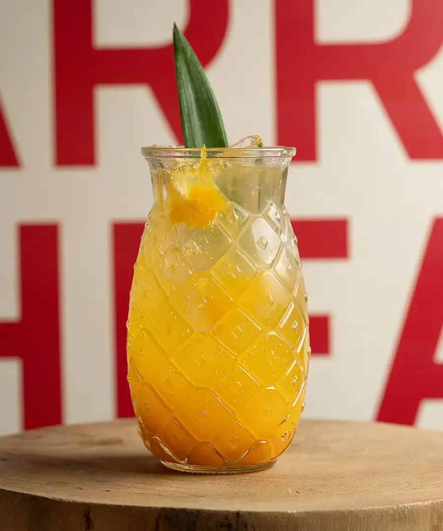 Cóctel de sabor tropical en una copa con forma de piña y decoración de rodaja de piña y hoja, en un fondo con letras rojas.