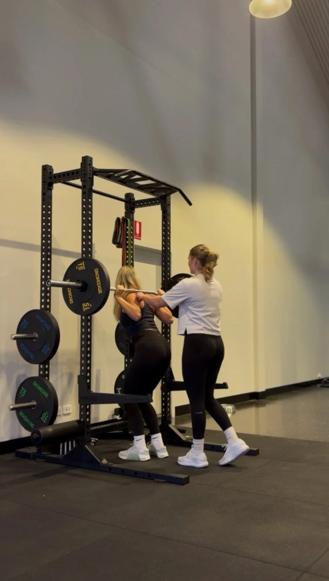 Two women are at a gym; one is spotting the other during a squat exercise with a barbell, while a trainer assists.