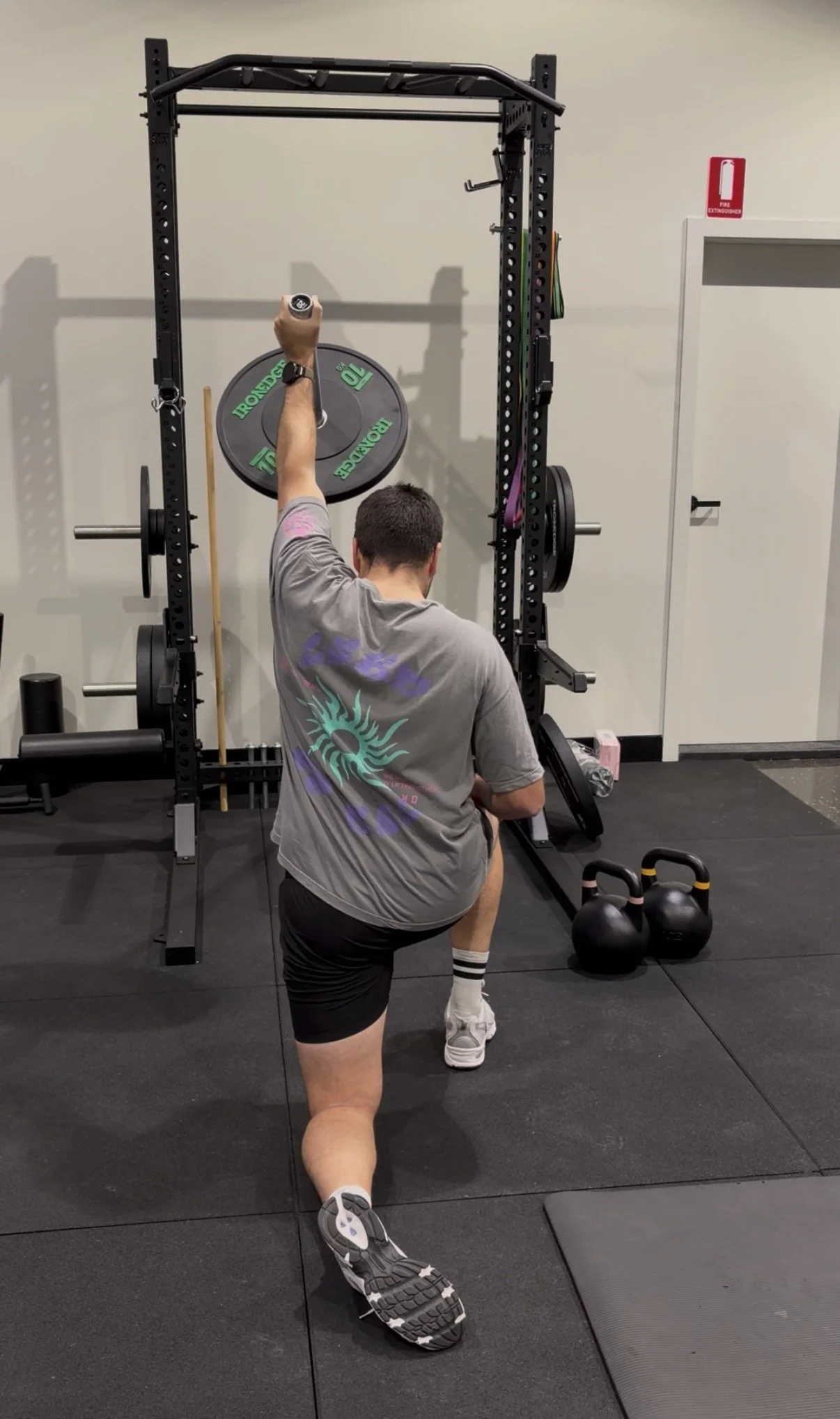 A man lifting a weight plate while kneeling on one knee in a gym, with kettlebells and workout equipment nearby.