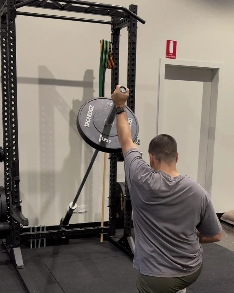 A man in a gym is performing a kneeling exercise with a barbell and a weight plate, in front of a squat rack. There are resistance bands hanging on the rack and a fire extinguisher on the wall.