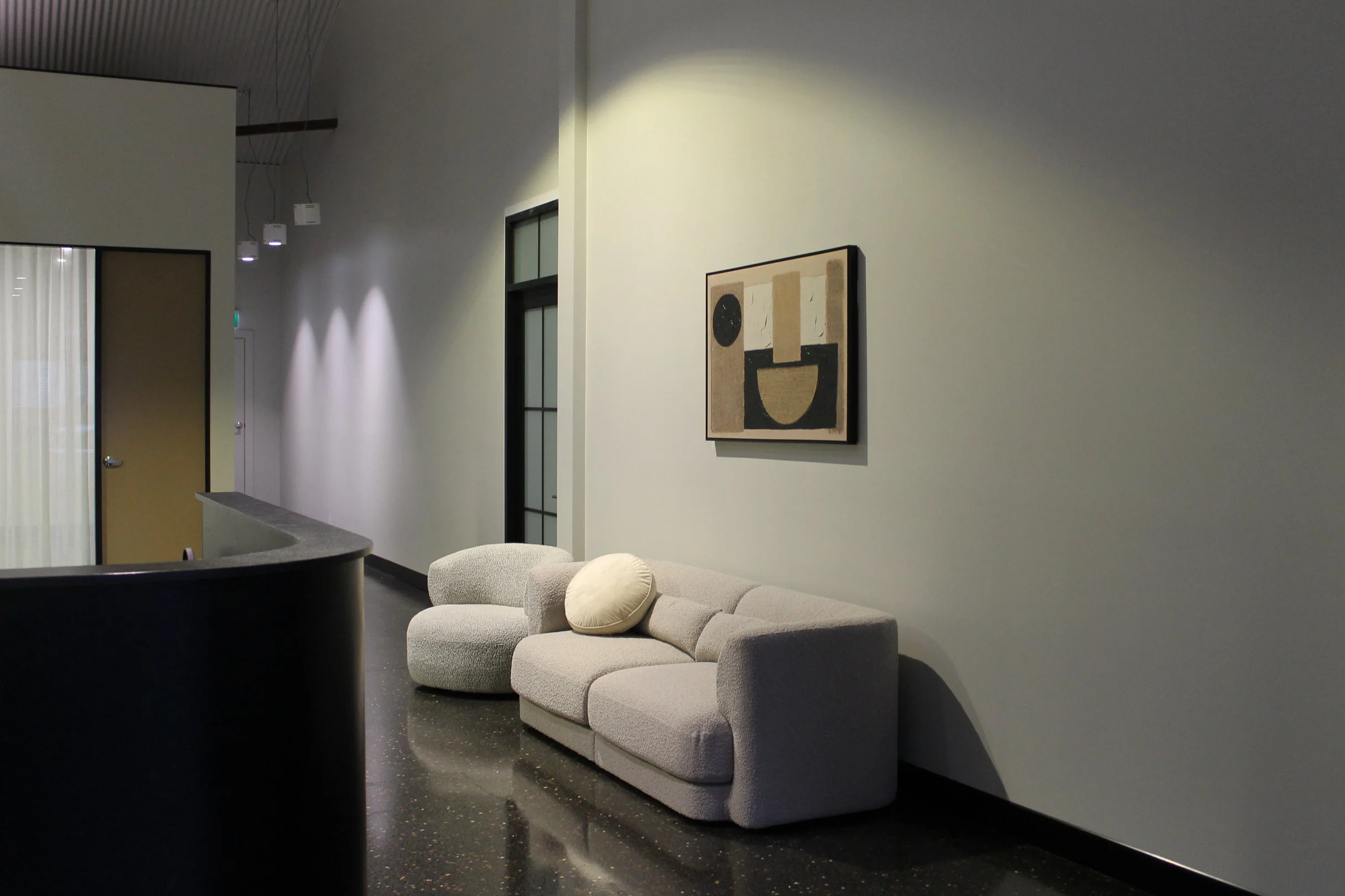 A modern interior space with grey walls, a curved grey sofa with round pillows, artwork on the wall, and lighting fixtures hanging from the ceiling.