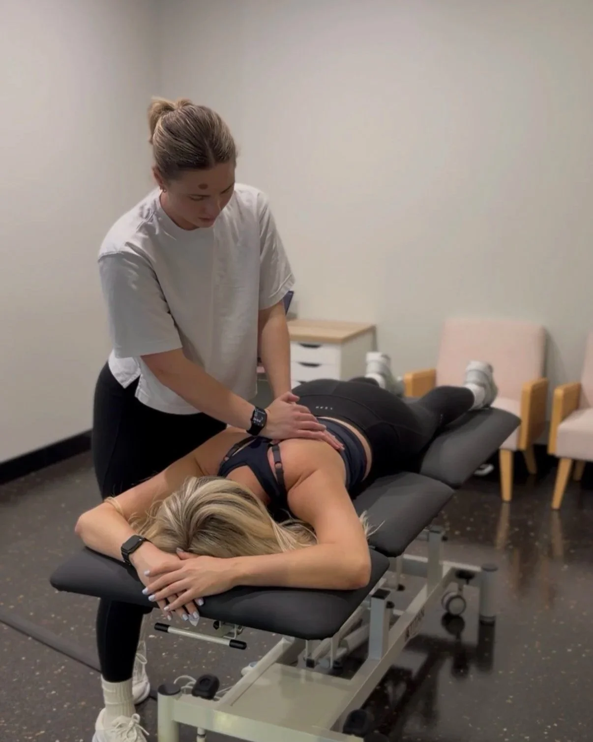 A woman receives chiropractic treatment on her back from a chiropractor in a clinical setting with chairs and a cabinet in the background.
