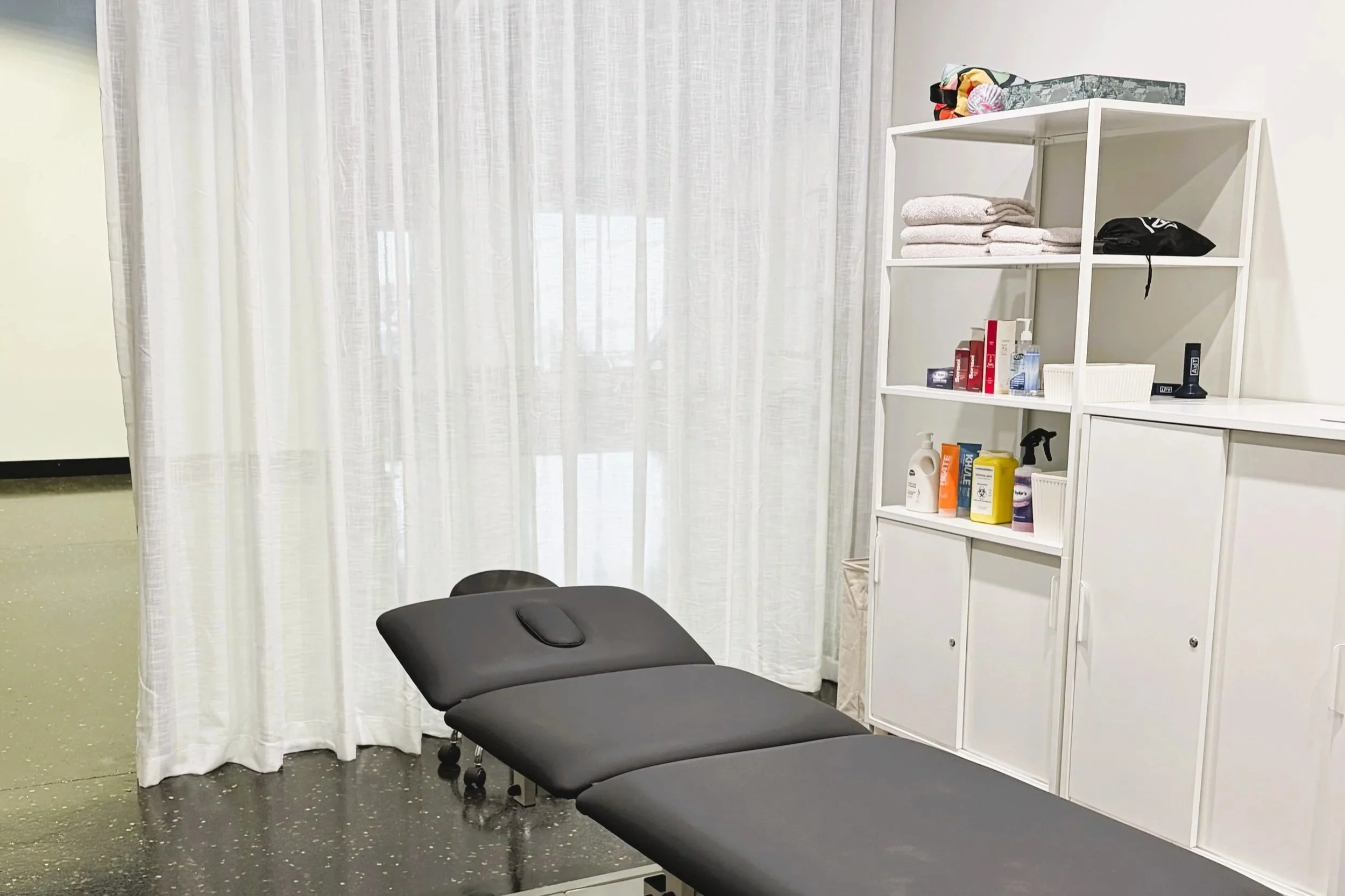 Medical examination room with black examination table, white storage shelf with towels and medical supplies, and white curtains.