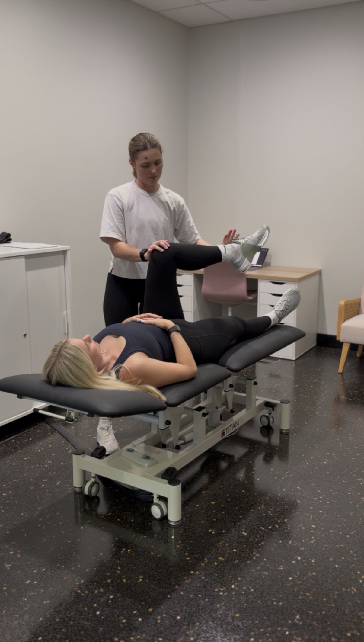 A woman lying on a medical examination table with her eyes closed, while a healthcare professional stands next to her holding her raised leg during a physical therapy session in a clinical room.