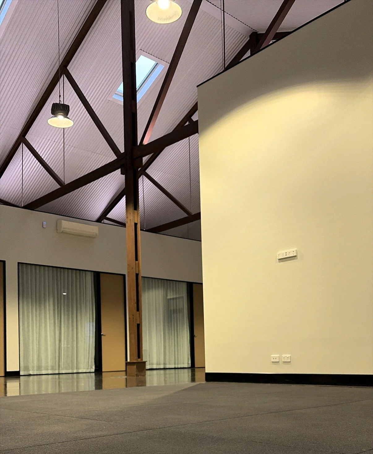 Interior of a modern building with a high, vaulted ceiling, exposed wooden beams, skylight, hanging pendant lights, a wall-mounted air conditioning unit, and large glass doors with vertical blinds.