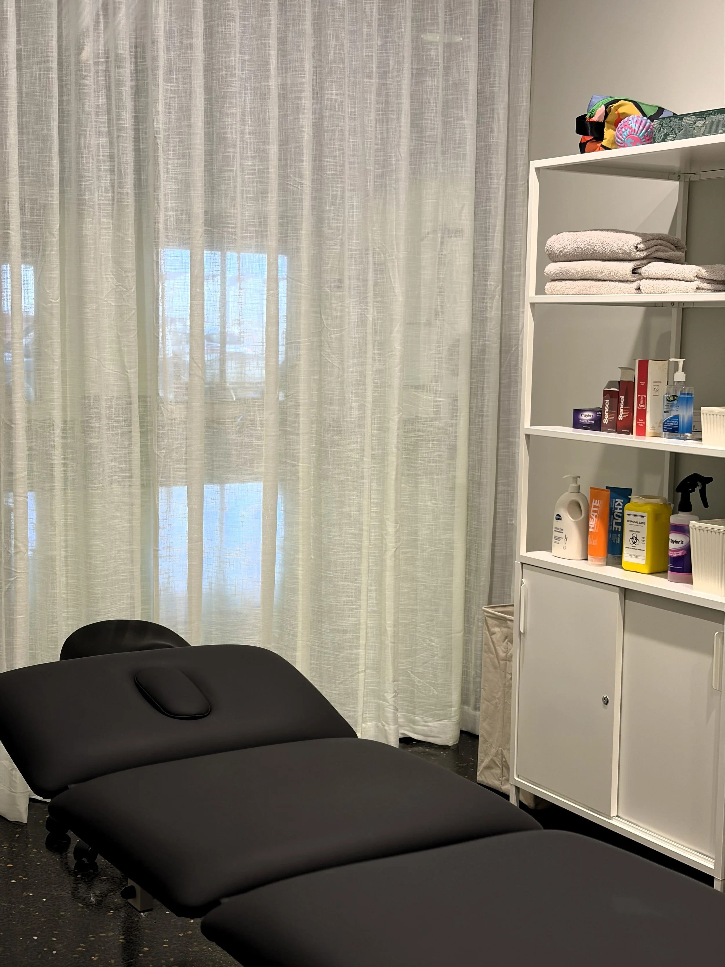 A black massage table in a room with a white storage cabinet, white curtains, and various personal care items on the shelves.