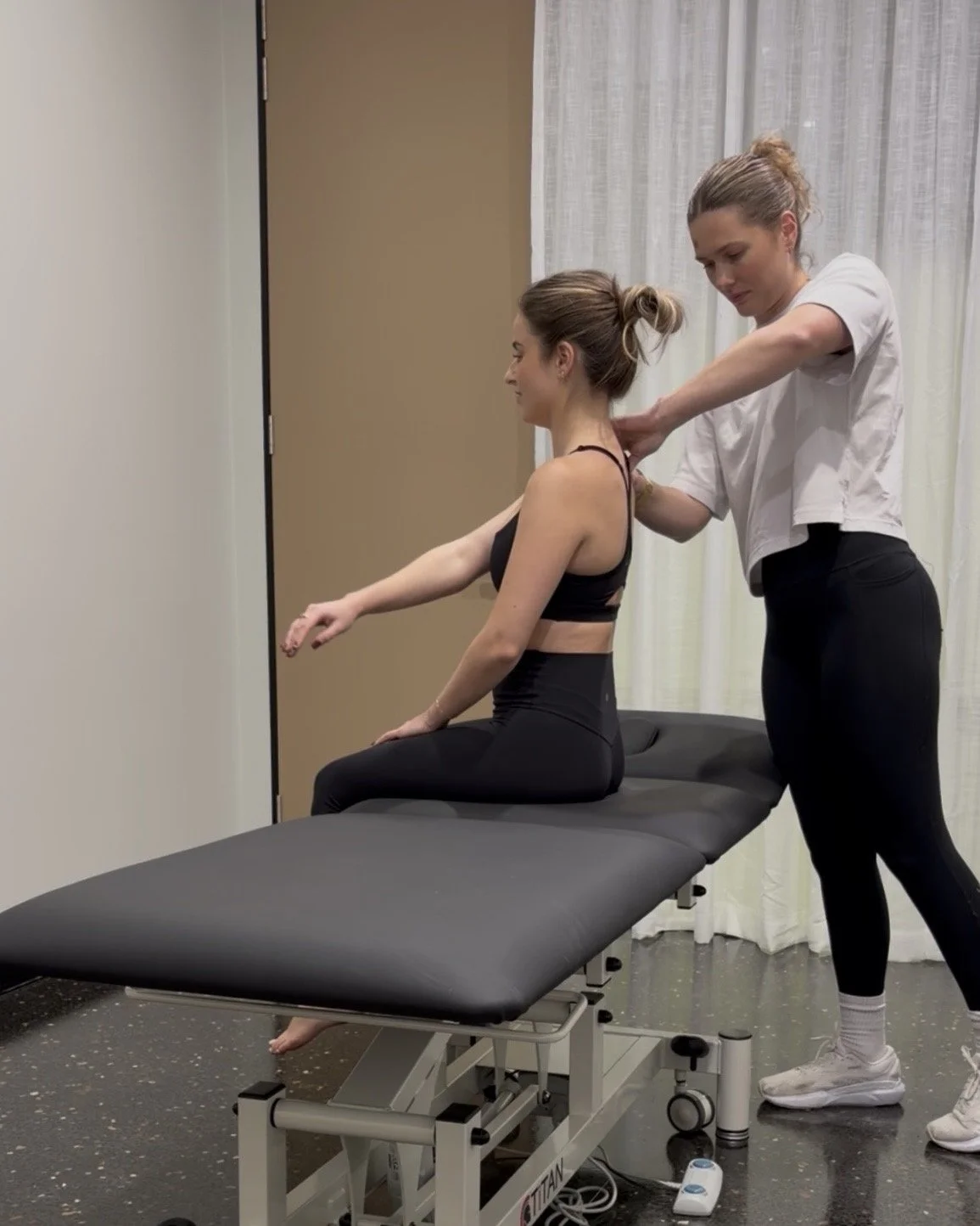A woman in black workout attire receives a neck and shoulder adjustment from a physiotherapist in a clinical setting with a massage table and white curtains.