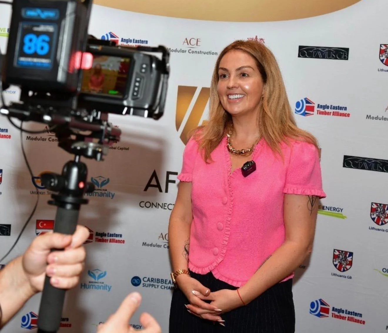 A woman with blonde hair wearing a pink top is being filmed or photographed at a backdrop with logos including Angus Eastern Timber Alliance, Humanity, and others.
