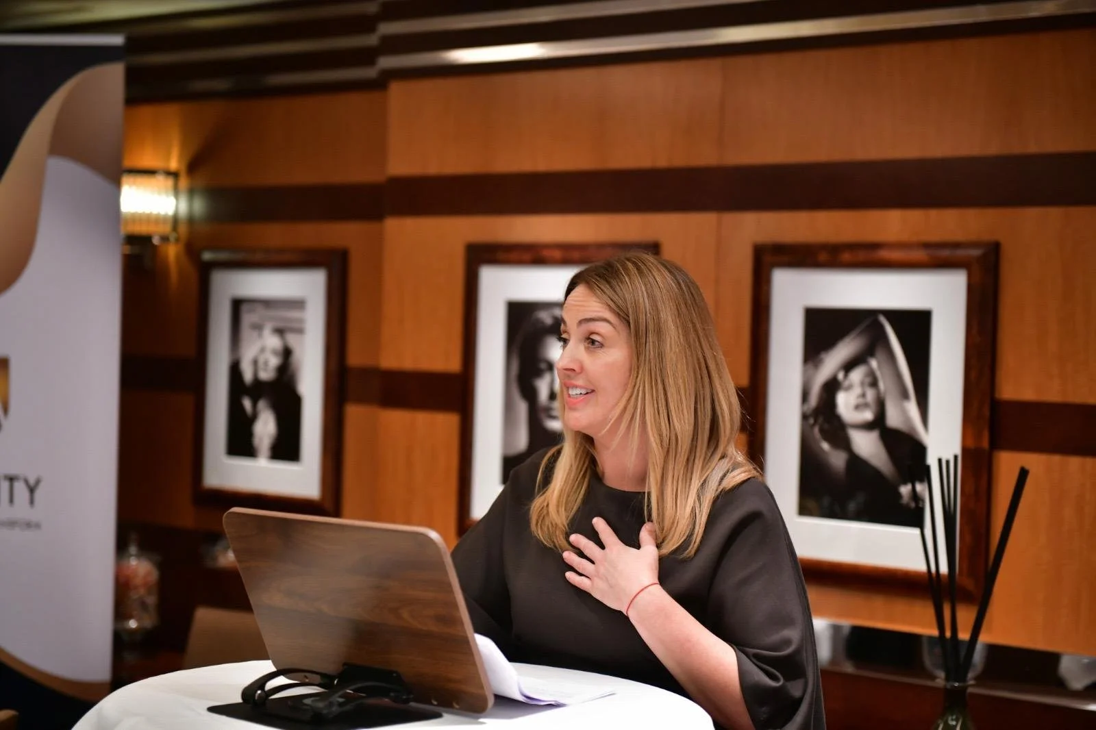 A woman with shoulder-length blonde hair, wearing a black top, sitting at a table with a laptop, gesturing with her right hand on her chest, in an art gallery or exhibition room with framed black and white photographs on the wooden wall behind her.