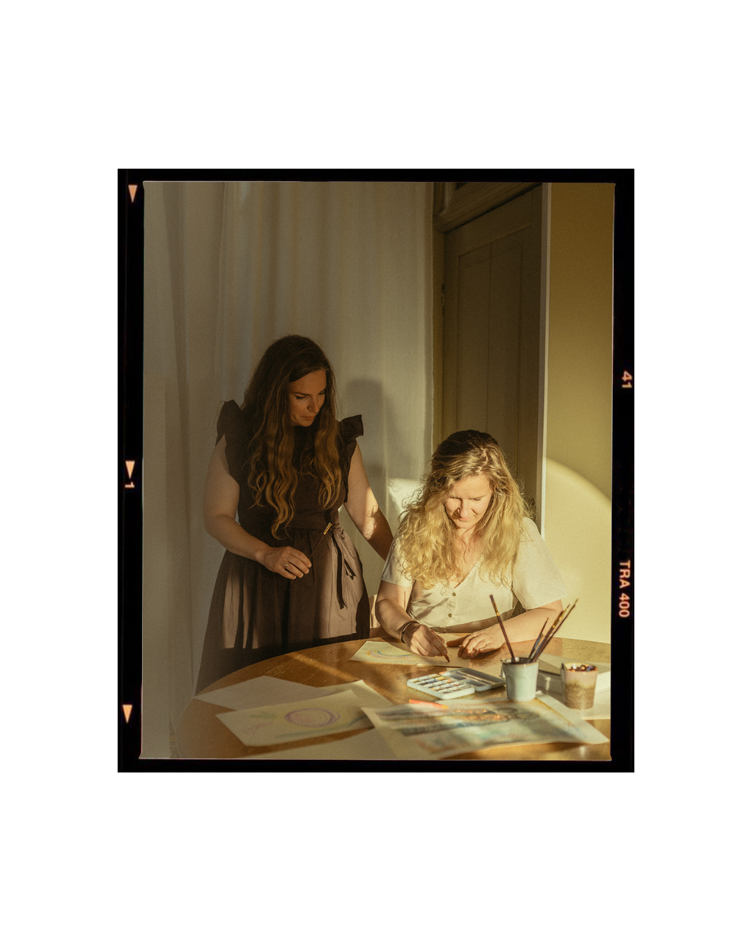 Twee vrouwen werken aan kunstprojecten aan een houten tafel met tekeningen, waterverf, en schildergereedschap.
