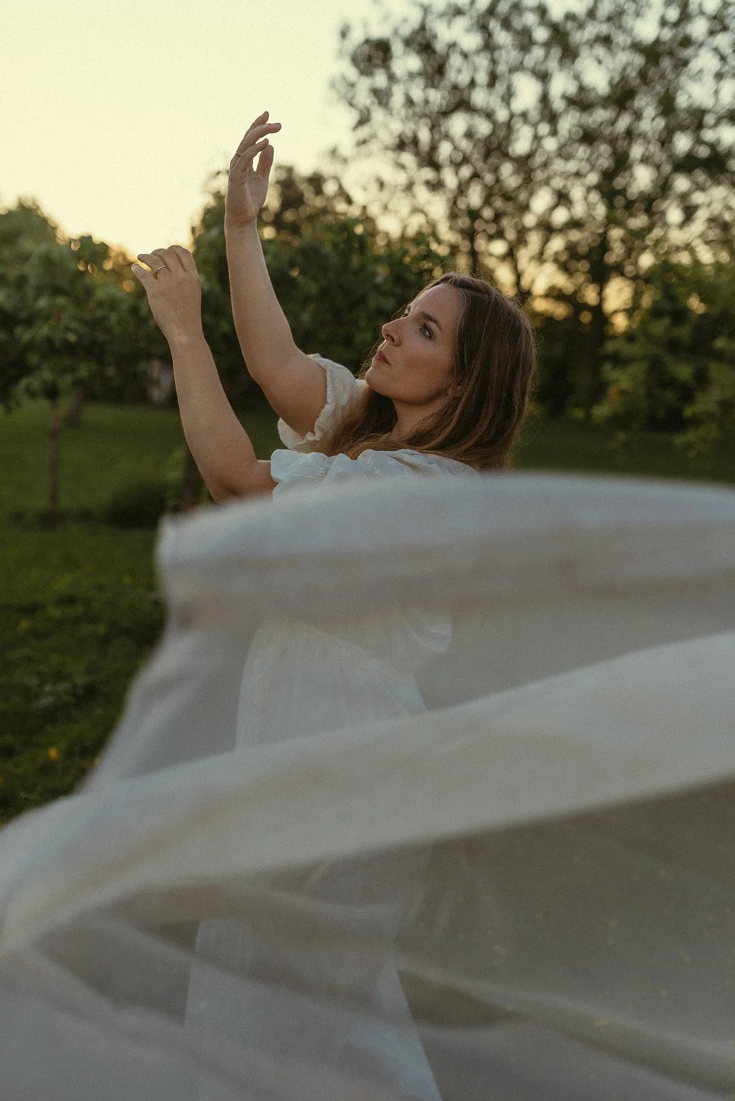 Een vrouw in een witte jurk die in een park poseert tijdens zonsondergang, met een boom op de achtergrond.