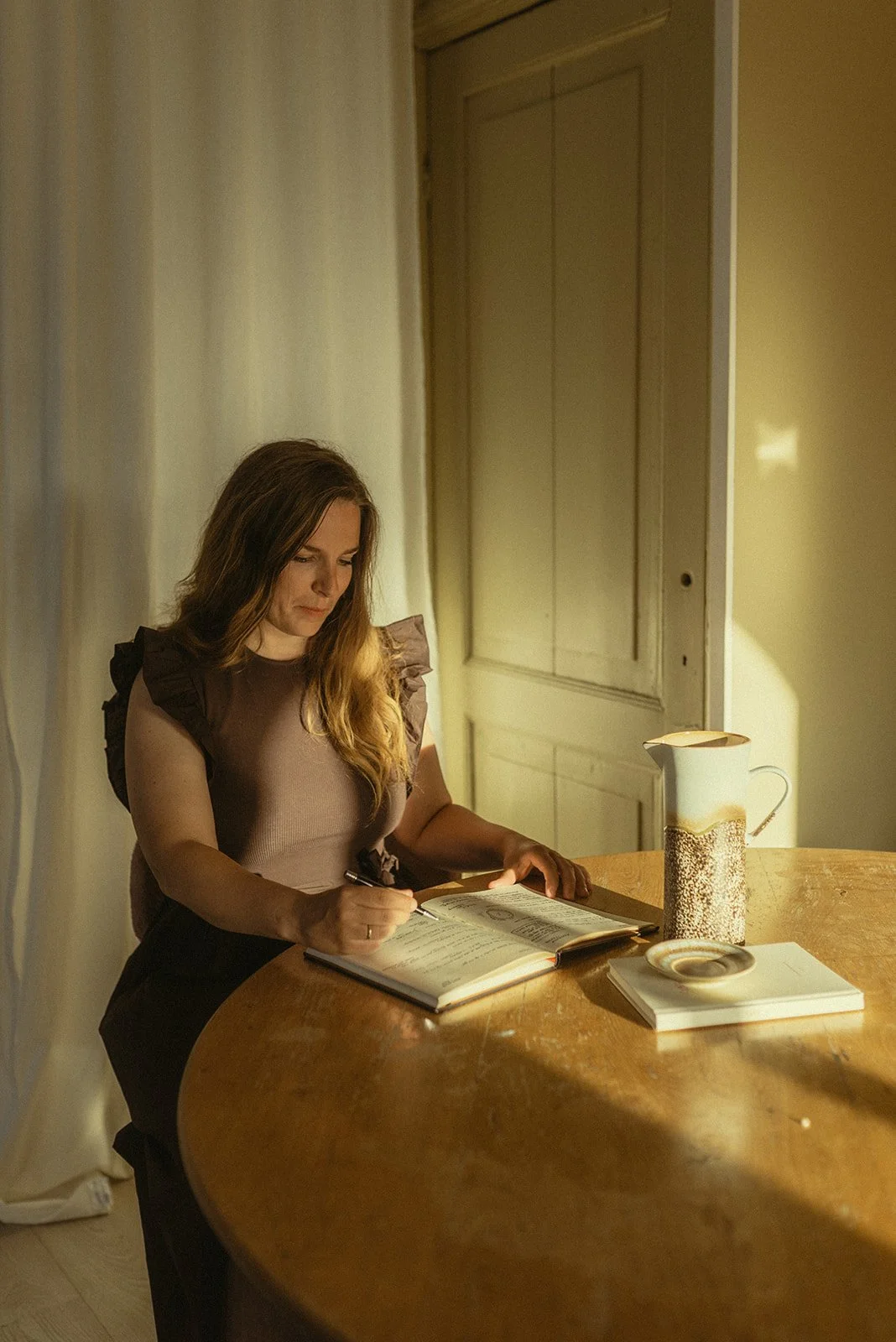 Vrouw schrijft in een notitieboekje aan een ronde houten tafel met een thermoskan, een kom en boeken.