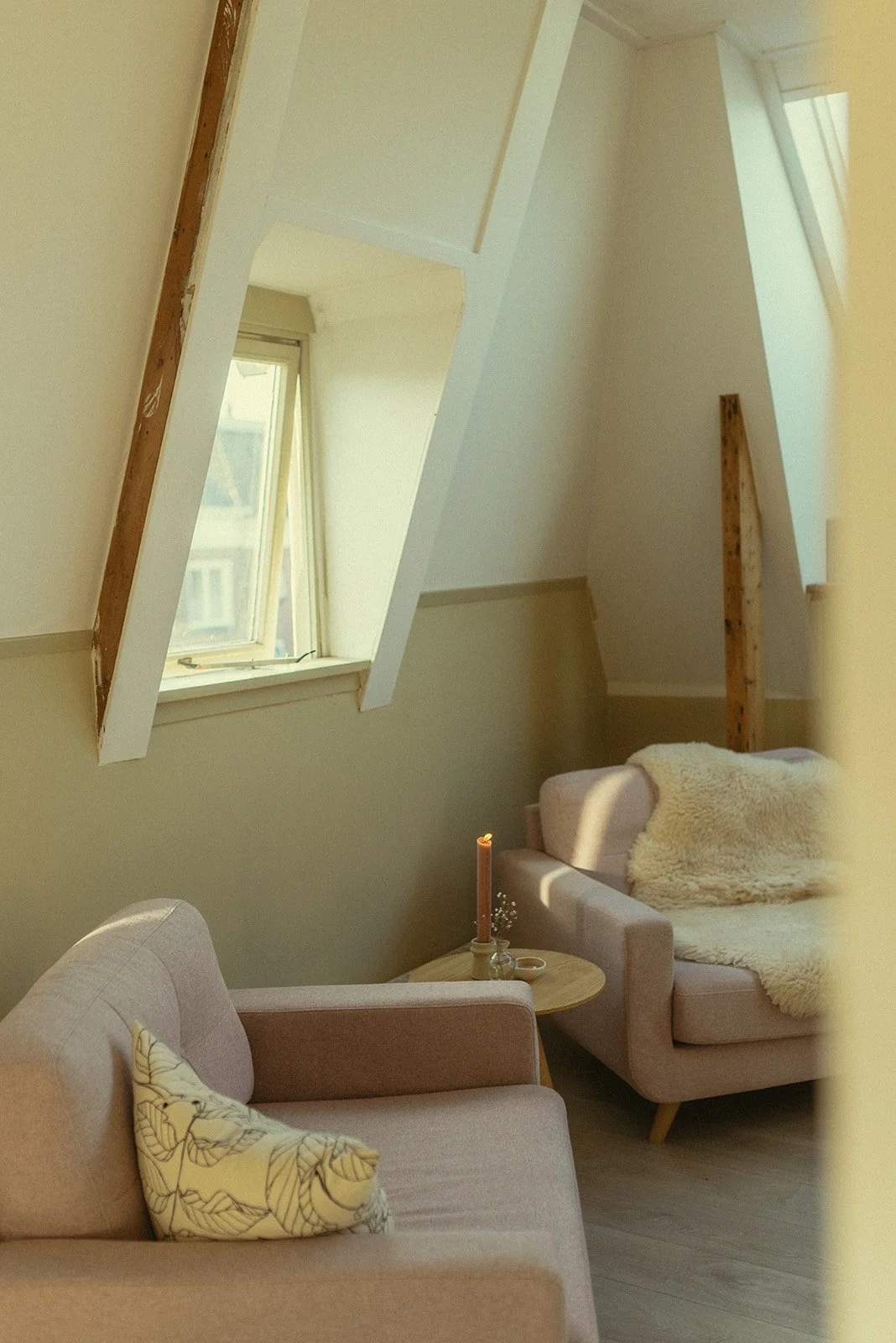 Een gezellige woonkamer met twee loungebanken, een kleine ronde tafel met een kaars, een lichtgele muur en een schuine dubbele beglazing venster.