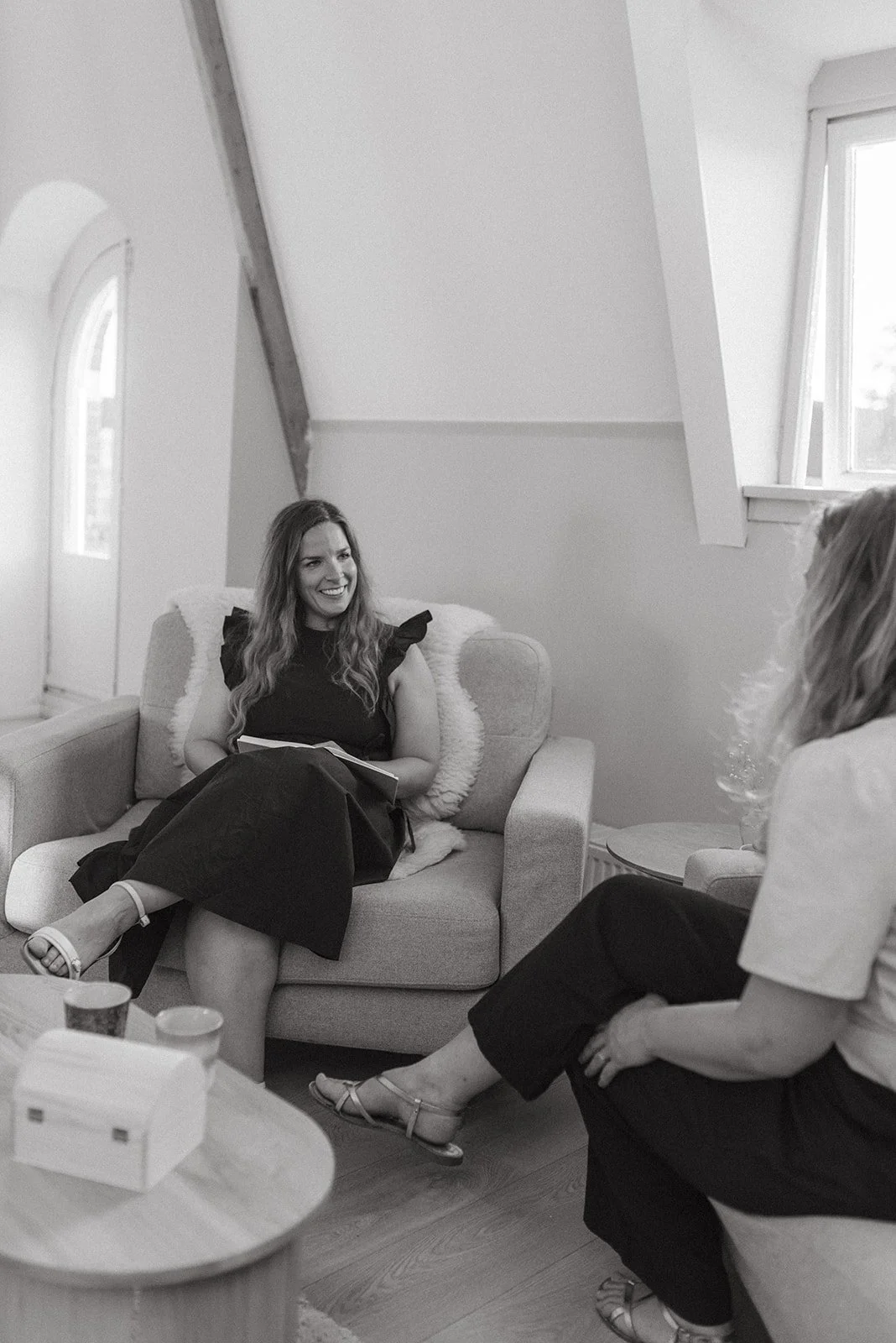 Twee vrouwen zitten in een woonkamer en hebben een gesprek. De ene vrouw zit op een bank en lacht, terwijl de andere vrouw luisterend zit.