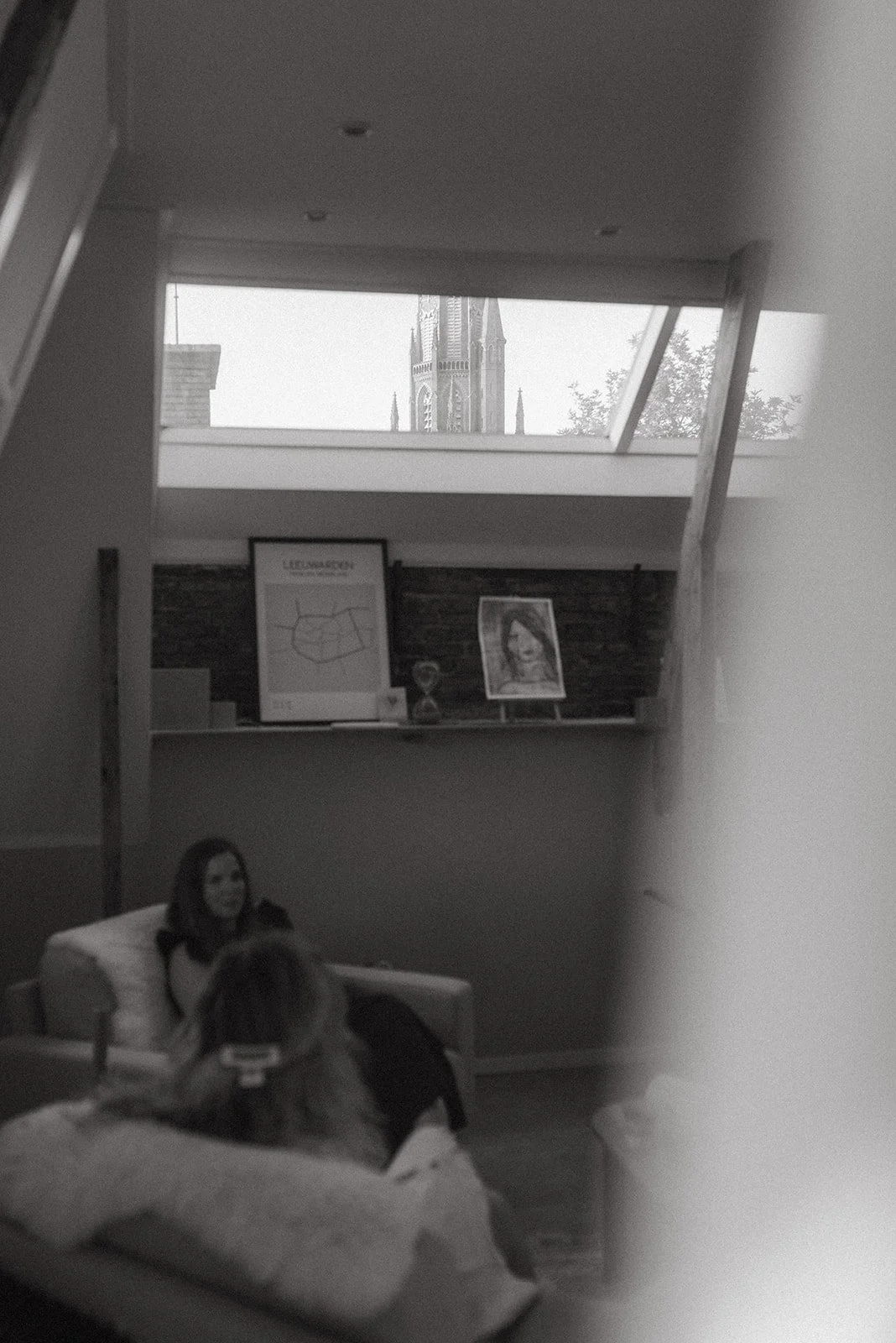 Interne ruimte met twee vrouwen, een zittend op een bank en een andere die haar met een smartphone filmt, binnen en een grote kerktoren zichtbaar door een raam boven.