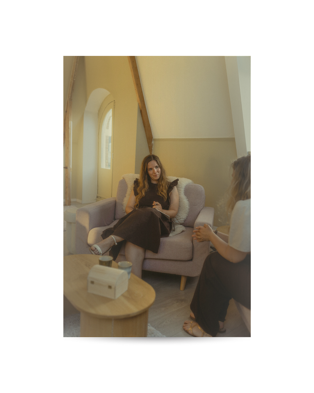 Twee vrouwen die een gesprek voeren in een gezellige woonkamer met een hoog plafond en grote ramen.