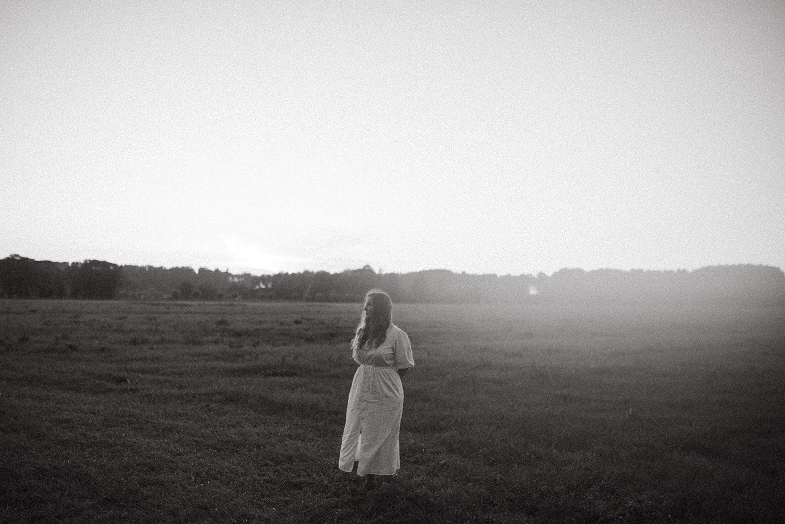 Een vrouw in een lange jurk staat op een open veld, met een silhouetachtig landschap op de achtergrond, alsof de zon net onder is gegaan of opkomt.