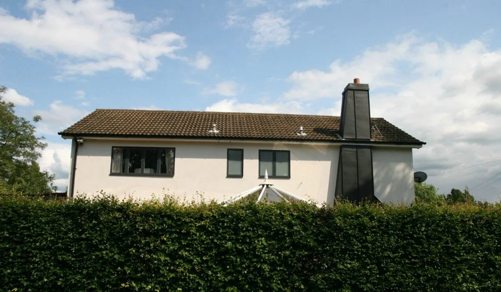 A two-story white house with a brown tiled roof, black framed windows, and a tall black chimney, surrounded by green bushes under a partly cloudy sky.
