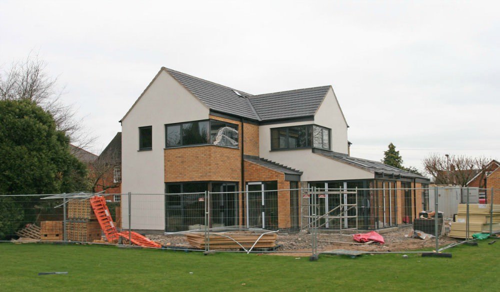 A modern two-story house under construction, fenced off with building materials and tools around, with a grassy yard in the foreground.