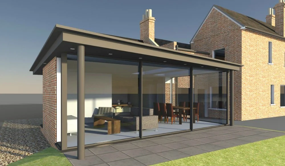 A modern backyard sunroom with glass walls and a flat roof, attached to a brick house with vaulted rooflines and chimneys, showing an interior with furniture including a sofa and dining table.