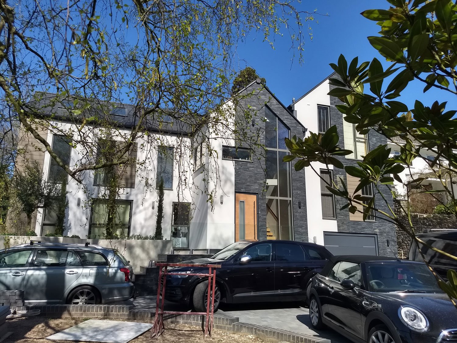Modern multi-story residential house with white walls, black accents, large glass windows, and a wooden front door, surrounded by trees with parked cars in front on a sunny day.