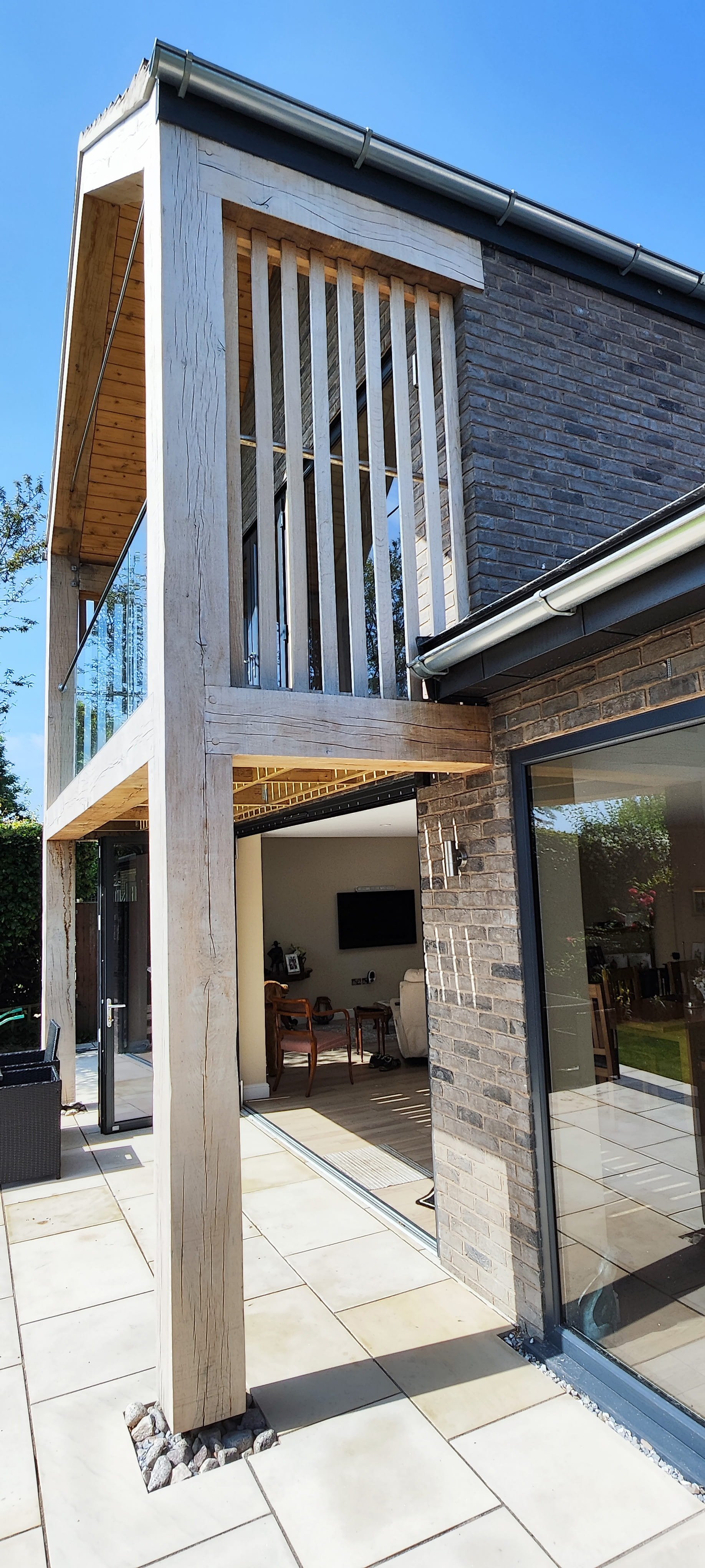 Modern house exterior with a patio, large sliding glass doors, brick walls, and a wooden balcony with vertical slats under a clear blue sky.