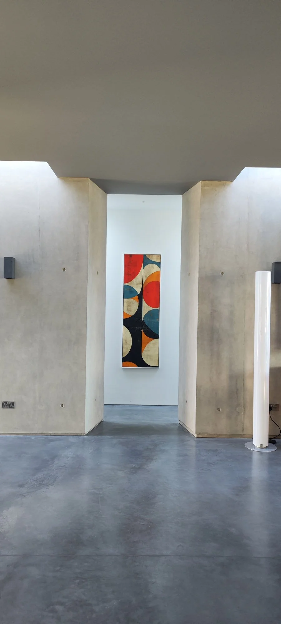 Modern art gallery with concrete walls and a colorful abstract painting on the back wall featuring circles and curves in red, blue, orange, beige, and black, with a polished concrete floor.
