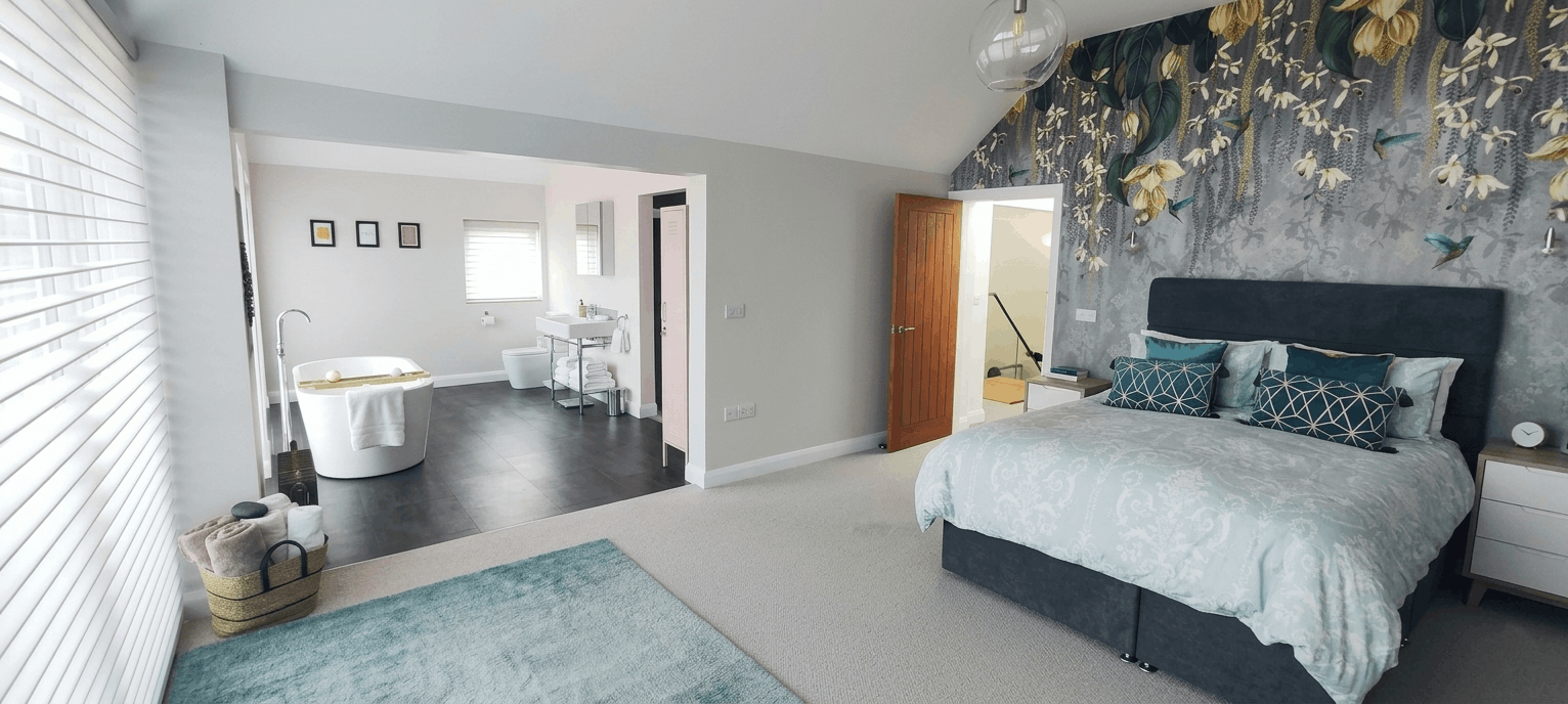 A spacious bedroom with a large bed featuring a dark headboard and multiple pillows, a pair of nightstands, and a decorative wallpaper with birds and plants. There is a bathroom area visible with a bathtub, sink, and toilet, separated by a partial wa