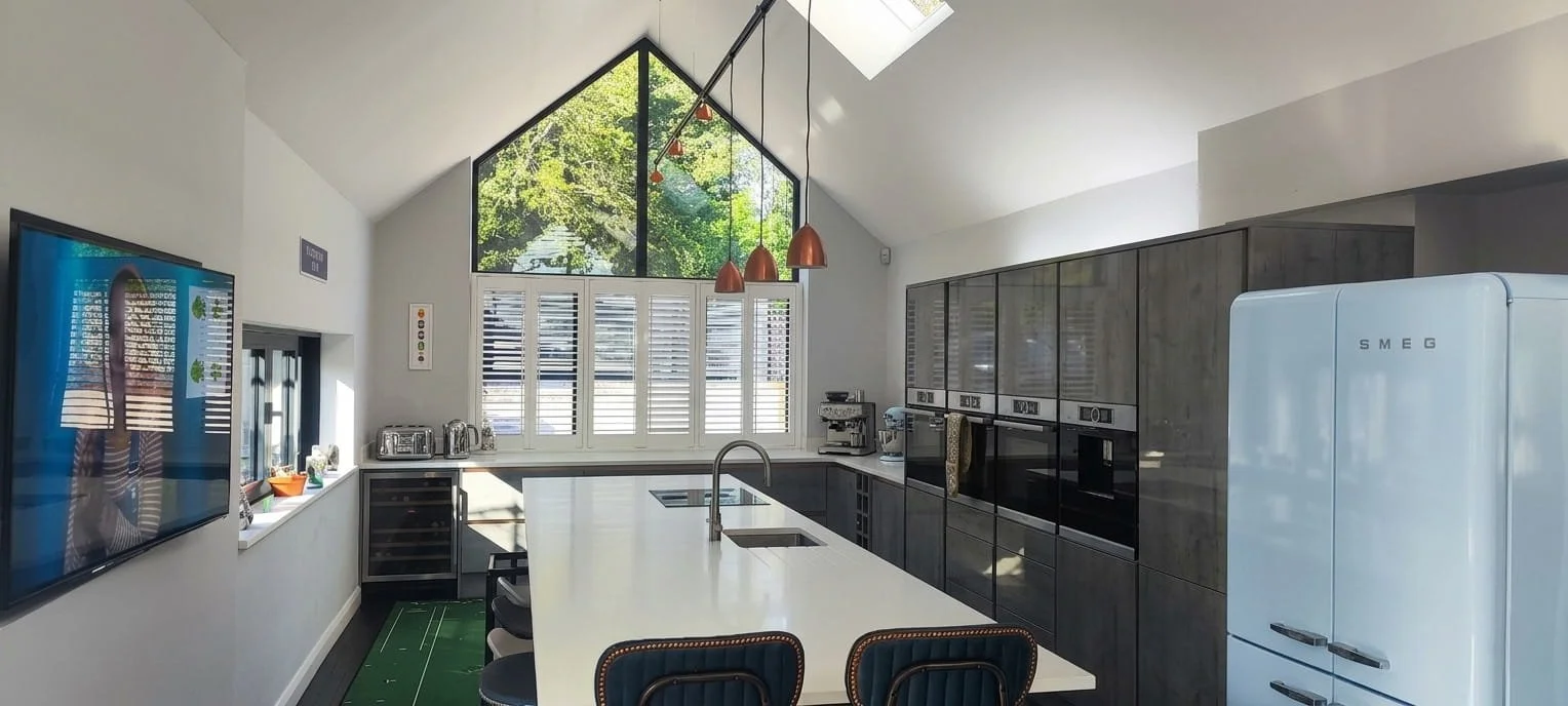 Modern kitchen with large central island, high vaulted ceiling, and big window showing green trees outside. Equipped with appliances like oven, microwave, and wine cooler, with bar stools at the island.